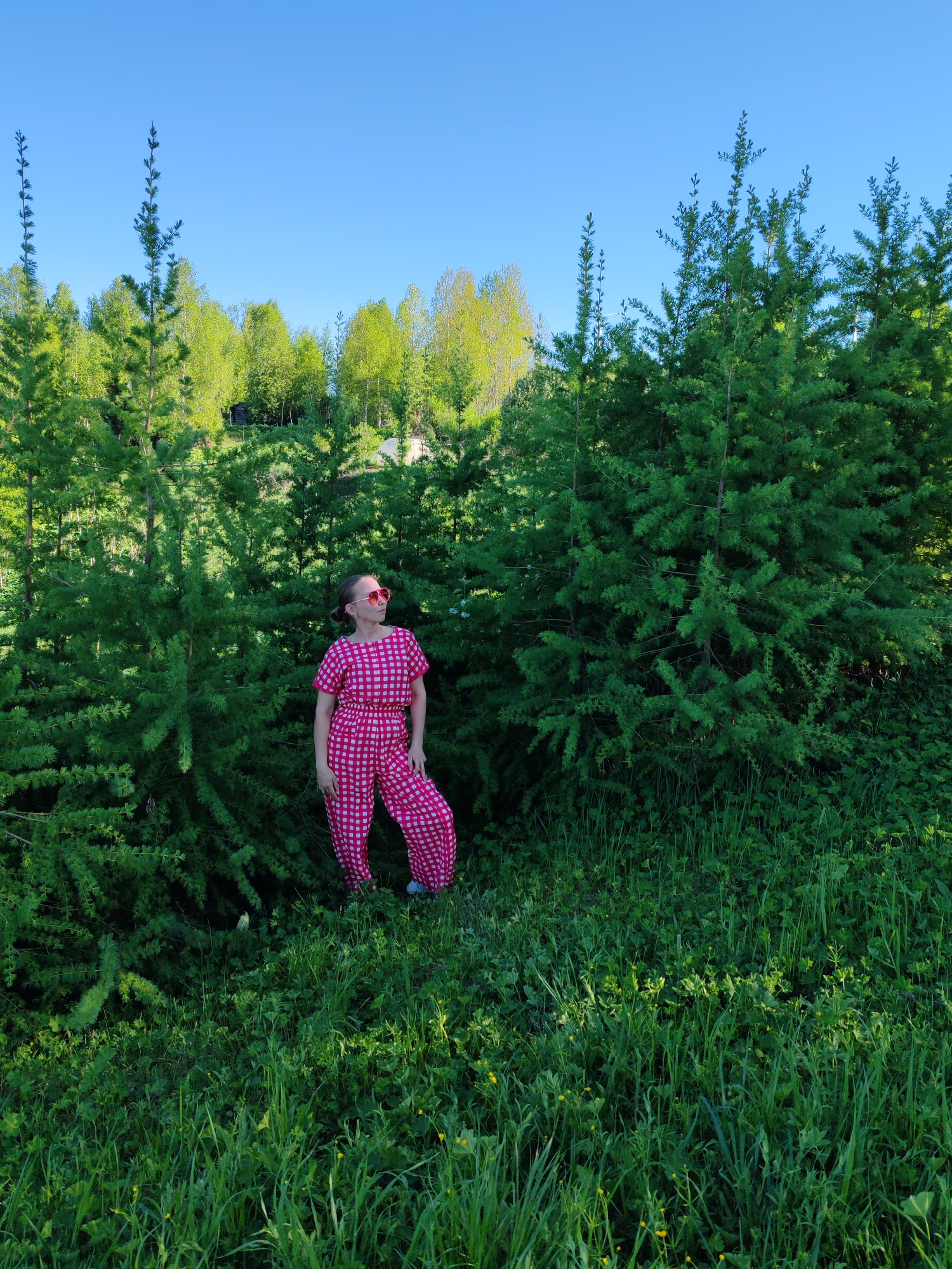 A woman stands in a grassy area in Чухлэм, Russia. She is wearing a pink and white checkered jumpsuit and pink sunglasses. She is surrounded by tall green coniferous trees. The grass is lush and green, with some small yellow flowers. The sky is a clear blue.