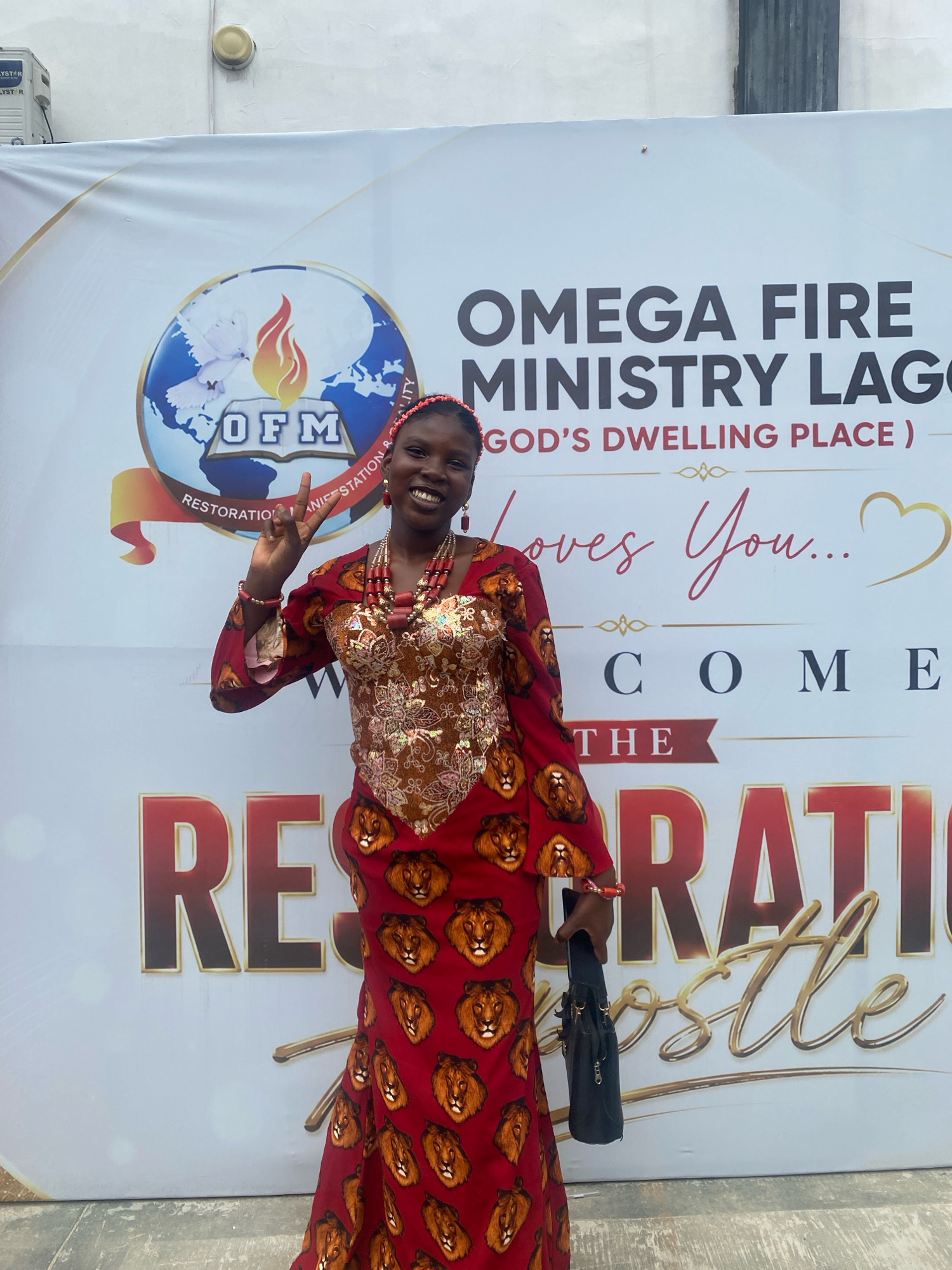 A smiling young African woman stands in front of a large white banner. The banner has text and logos for "OMEGA FIRE MINISTRY LAGOS" and "GOD'S DWELLING PLACE". The woman is wearing a red dress with a lion print, layered necklaces, earrings, and a red headband. She is holding a black purse in her right hand and has her left hand raised in a peace sign. The background appears to be an outdoor setting, possibly in Ado, Nigeria, given the ministry's name. The lighting suggests it is daytime.