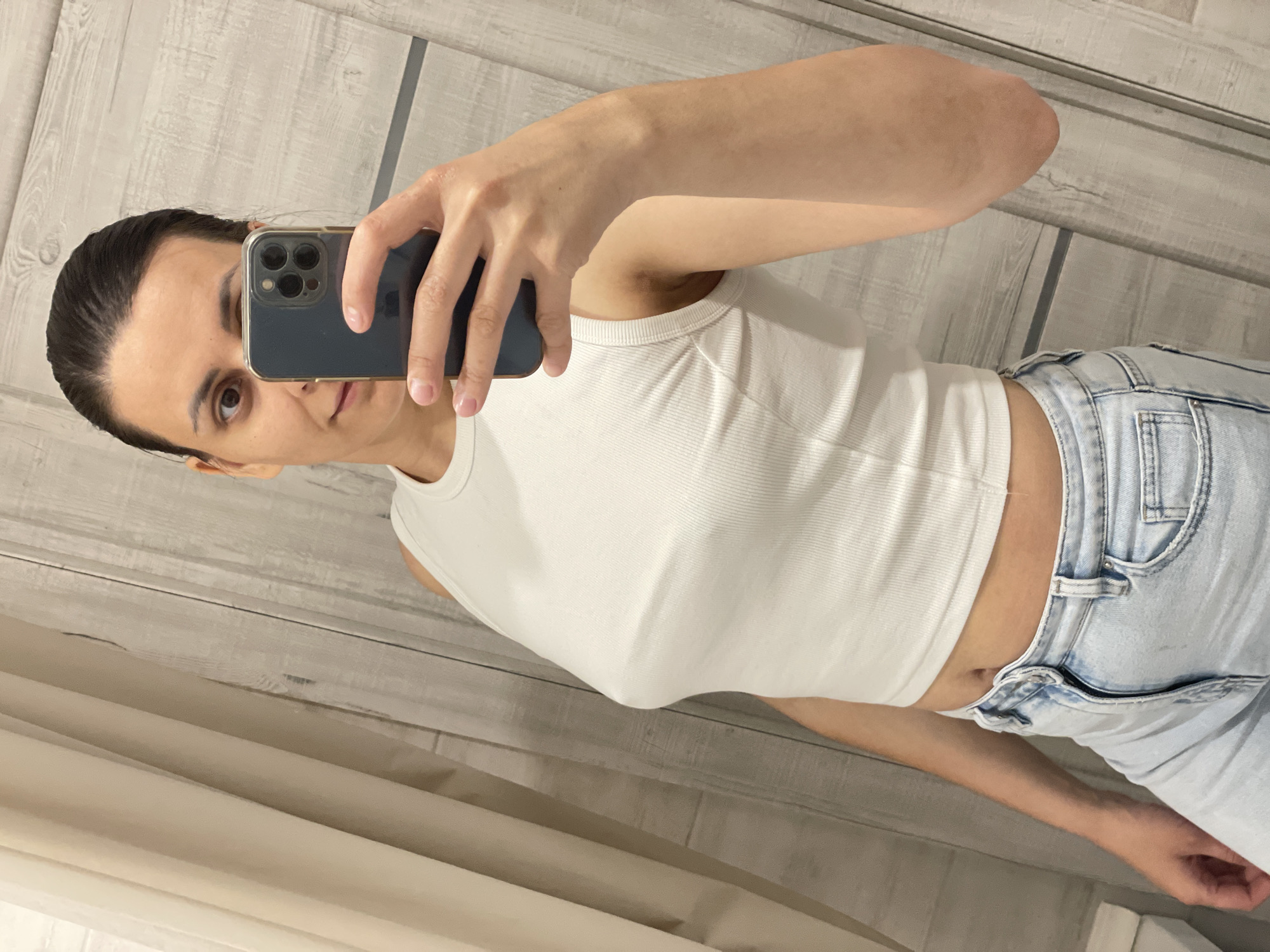 A woman is taking a selfie in an indoor location. She is holding a dark-colored smartphone in her right hand. She has dark hair pulled back and is wearing a white ribbed tank top and light blue jeans. The background appears to be a light-colored, possibly wood-paneled wall. The location is Unknown, Unknown.