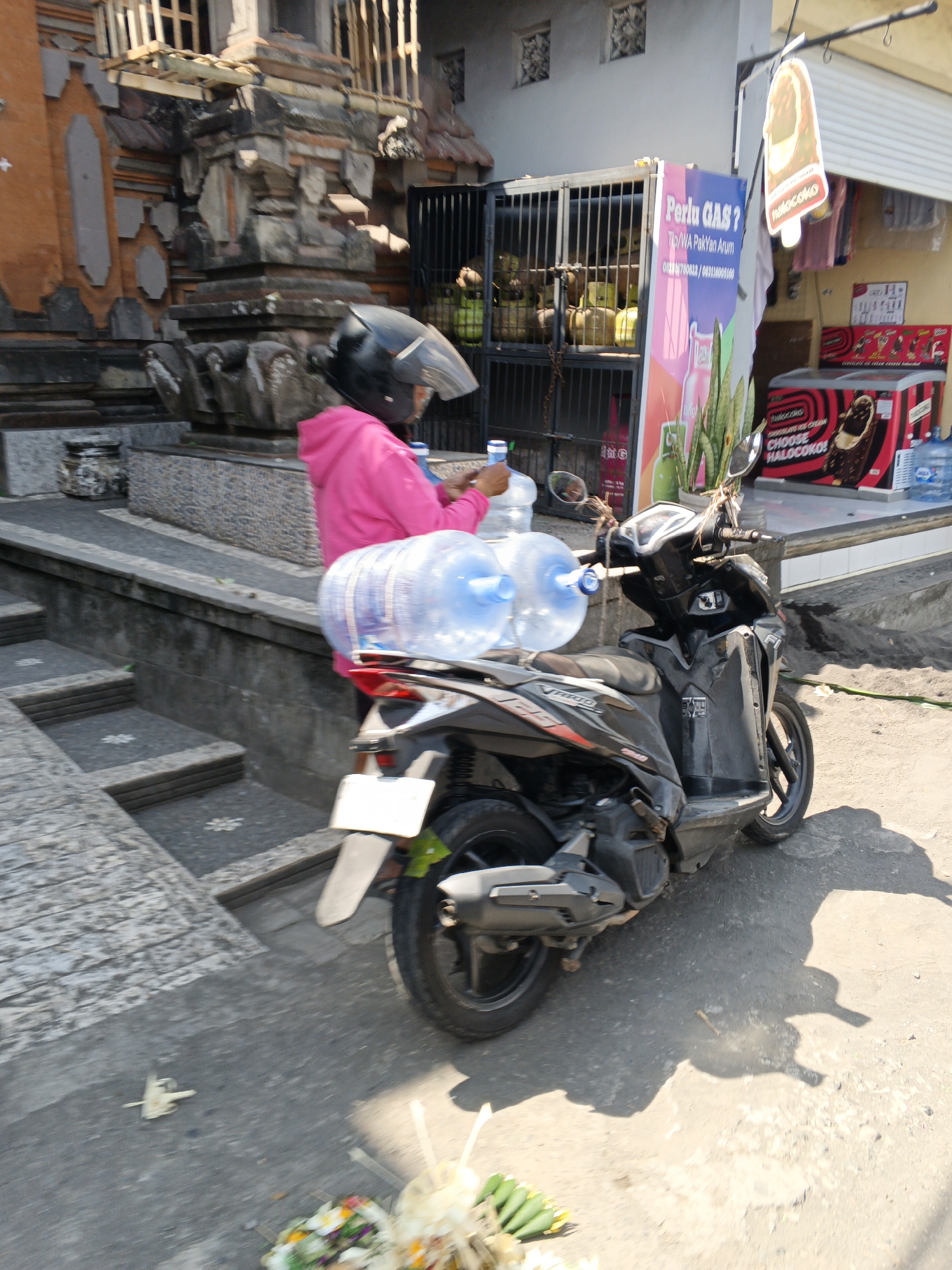 In the district of Ubud, Indonesia, a person wearing a bright pink hooded jacket and a black helmet is positioned behind a black scooter. The scooter is carrying two large, clear plastic water jugs strapped to its rear. The jugs are filled with a light blue liquid. The scooter is parked on a paved surface next to a set of stone stairs. In the background, there is a building constructed with stone and orange plaster, featuring decorative architectural elements and a barred gate behind which some green objects, possibly coconuts, are visible. Adjacent to this building is a shopfront displaying promotional posters for "Perlu GAS?" and "Halococo" ice cream, with a red refrigerator unit visible. The scene is bathed in bright sunlight, casting shadows on the ground.