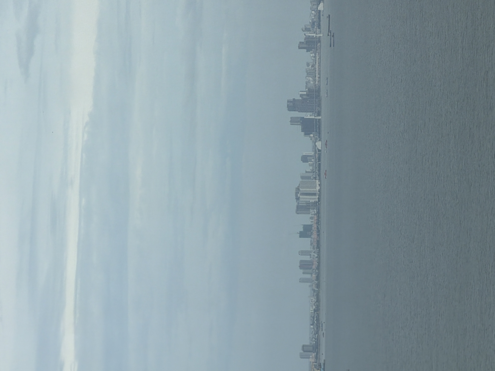 This is a wide-angle, aerial shot of a sprawling city skyline situated across a body of water. The city, identified as Hải Vân Ward, Vietnam, features a dense collection of high-rise buildings, including skyscrapers, apartment complexes, and what appear to be under-construction towers. The buildings are varied in height and architectural style, with some being modern glass structures and others more traditional.
The body of water in the foreground is calm and appears to be a bay or a harbor, with several vessels visible. These include cargo ships, smaller boats, and potentially ferries, dotting the surface of the water.
The sky above is overcast, with layers of clouds creating a muted, diffused light that casts a soft glow over the scene. The overall atmosphere is serene, with the city lights subtly visible in the distance, suggesting it is either early morning or late afternoon. The horizon line is sharp, separating the cityscape from the expanse of water and sky. There are no visible people in the frame, and the focus is on the urban landscape and its relationship with the surrounding environment.