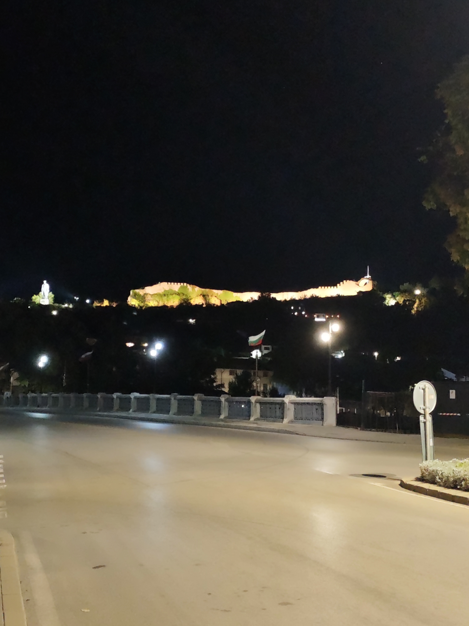 The image shows a nighttime view of the fortress of Lovech, Bulgaria, illuminated against the dark sky. The fortress, with its characteristic long, elevated structure, is prominently lit with warm yellow lights, highlighting its historical architecture. In the foreground, a street or road is visible, appearing wet and reflecting the ambient light, suggesting it might have rained. A stone barrier or retaining wall lines the edge of the road. Several streetlights are also visible, casting pools of light on the road and surrounding areas. A Bulgarian flag is seen flying on a flagpole in the middle ground. The overall atmosphere is serene and picturesque, with the historical landmark serving as the focal point.