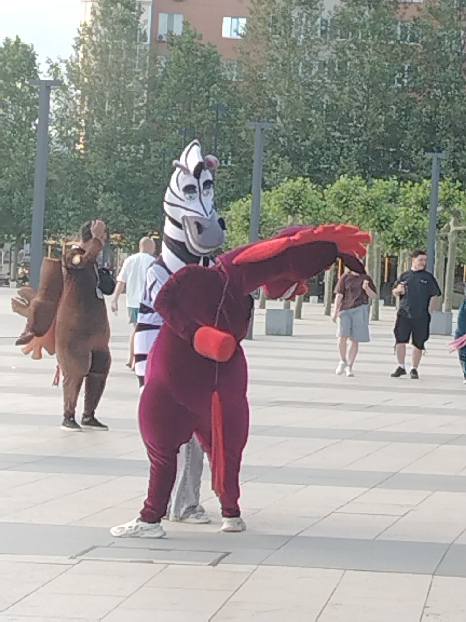 An outdoor shot shows people in mascot costumes and regular clothing in what appears to be a public plaza. A person in a zebra costume with black and white stripes and pink ears stands front and center, facing right and holding their arms out. To the left of the zebra, a person in a brown bird-like costume with large wings stands facing left. Behind the zebra, another person in a maroon costume with a large, feathered headpiece is partially visible. Other individuals in casual clothing are dispersed throughout the background, some walking and others standing. The environment features a paved ground with a grey and white tiled pattern, stone bollards, and trees lining the background, with a modern building visible beyond the trees. The lighting suggests it is daytime, possibly late afternoon. The scene is likely in an urban setting, possibly a park or public square in Krasnodar, Russia, given the architectural style of the building and the types of trees.