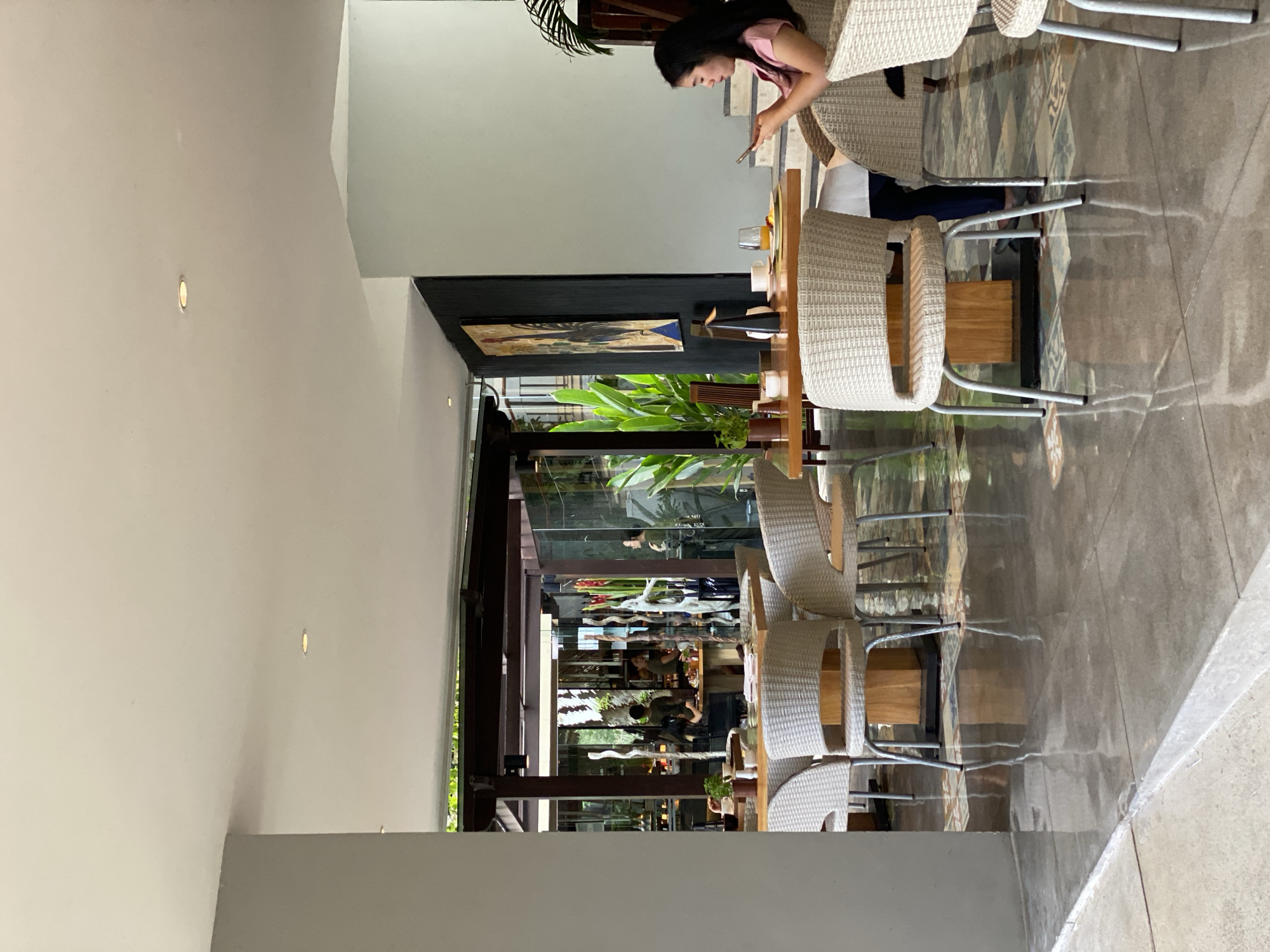 The image depicts an indoor dining area of a restaurant in Kuta Selatan, Indonesia. The setting features several tables and chairs, with a woman in a pink shirt sitting at one table in the foreground, looking at her phone. The tables are made of wood, and the chairs are woven with a light-colored material. Beyond the immediate seating area, a glass-enclosed section reveals more diners and staff in what appears to be a kitchen or service area. Lush green plants are visible both inside and outside the restaurant, contributing to a tropical ambiance. The floor is a combination of polished concrete and decorative tiles. The lighting suggests it is daytime, with natural light filtering in through the open and glassed areas, supplemented by artificial lighting from ceiling fixtures. A large painting of a rooster is displayed on a dark wall in the background. The overall impression is of a modern, airy, and somewhat upscale dining establishment.