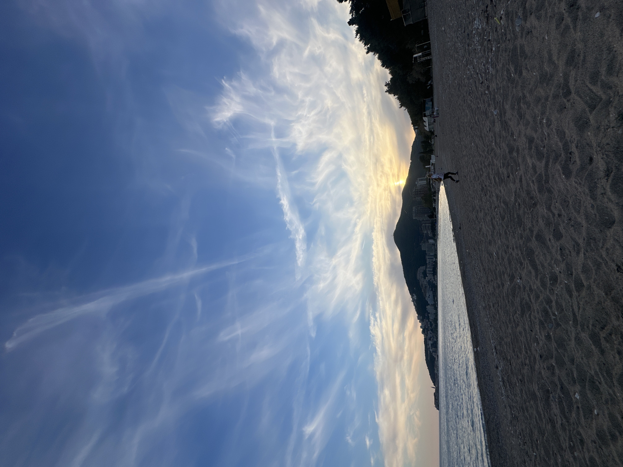 The image captures a beach scene in Budva, Montenegro at dawn. The sky is light blue with white cirrus clouds spreading vertically. The sun rises behind a mountain, casting a golden glow through the clouds. The beach is dark sand, and a person stands near the shoreline. Buildings are visible at the base of the mountain, near the beach's edge. The sea is calm.