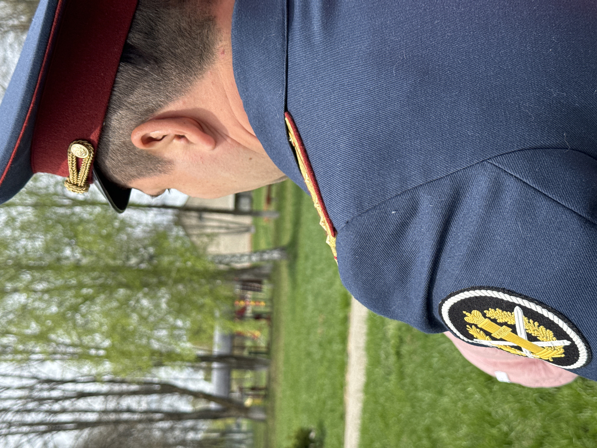 The image depicts a person in a military uniform, including a blue jacket with a patch featuring a sword and wheat stalks, and a hat with a red band and gold embellishments. The person is outside in a grassy area with trees and a path visible in the background. The location may be a cemetery or memorial site, suggested by the presence of grave markers or monuments. Based on the information provided, the location is within the city of Красный Маяк, Russia.