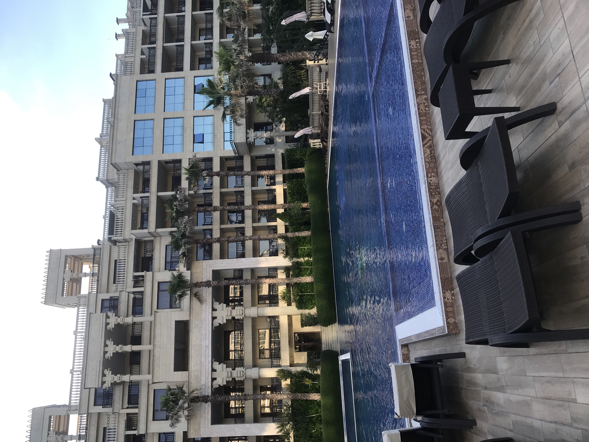 The image shows a swimming pool area in front of a large, multi-story hotel with an architectural style reminiscent of ancient Egyptian influences, featuring pillars with ornate capitals and stepped rooflines. The hotel is built of light-colored stone and has many balconies with dark railings. Palm trees are visible in front of the hotel. The swimming pool is rectangular with blue tiling. Around the pool, there are dark-colored lounge chairs and a small table. In the background, several closed white umbrellas are positioned along a railing. The scene appears to be in the city of Varna, Bulgaria, given the hotel's distinctive design. The sky is clear and bright, suggesting daytime.
