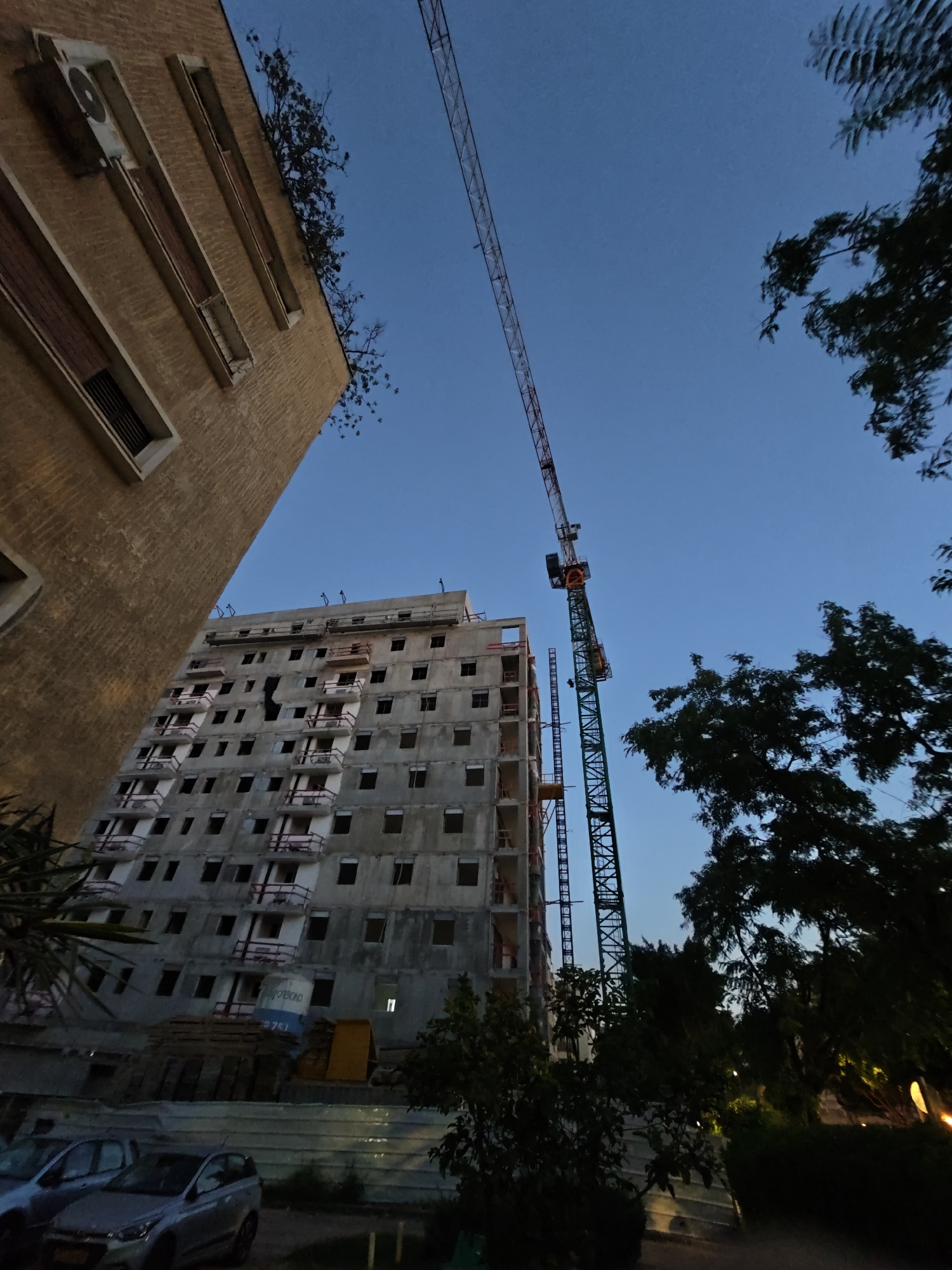 The image depicts an outdoor construction site in Petah Tikva, Israel, at dusk. A tall, green tower crane is prominently positioned, with its red boom extending towards the clear, blue sky. Adjacent to the crane is a multi-story building under construction, characterized by its unfinished concrete facade and numerous open window frames. Some balconies are visible, partially constructed with red railings.
To the left of the construction site, a portion of a beige brick building is visible, featuring several horizontal window openings. An air conditioning unit is mounted on this building. Trees with dark green foliage are scattered throughout the scene, particularly on the right side, framing the construction activity. In the foreground, two cars are parked, one silver and one a lighter shade, on what appears to be a paved surface. The overall atmosphere suggests ongoing development in an urban environment.