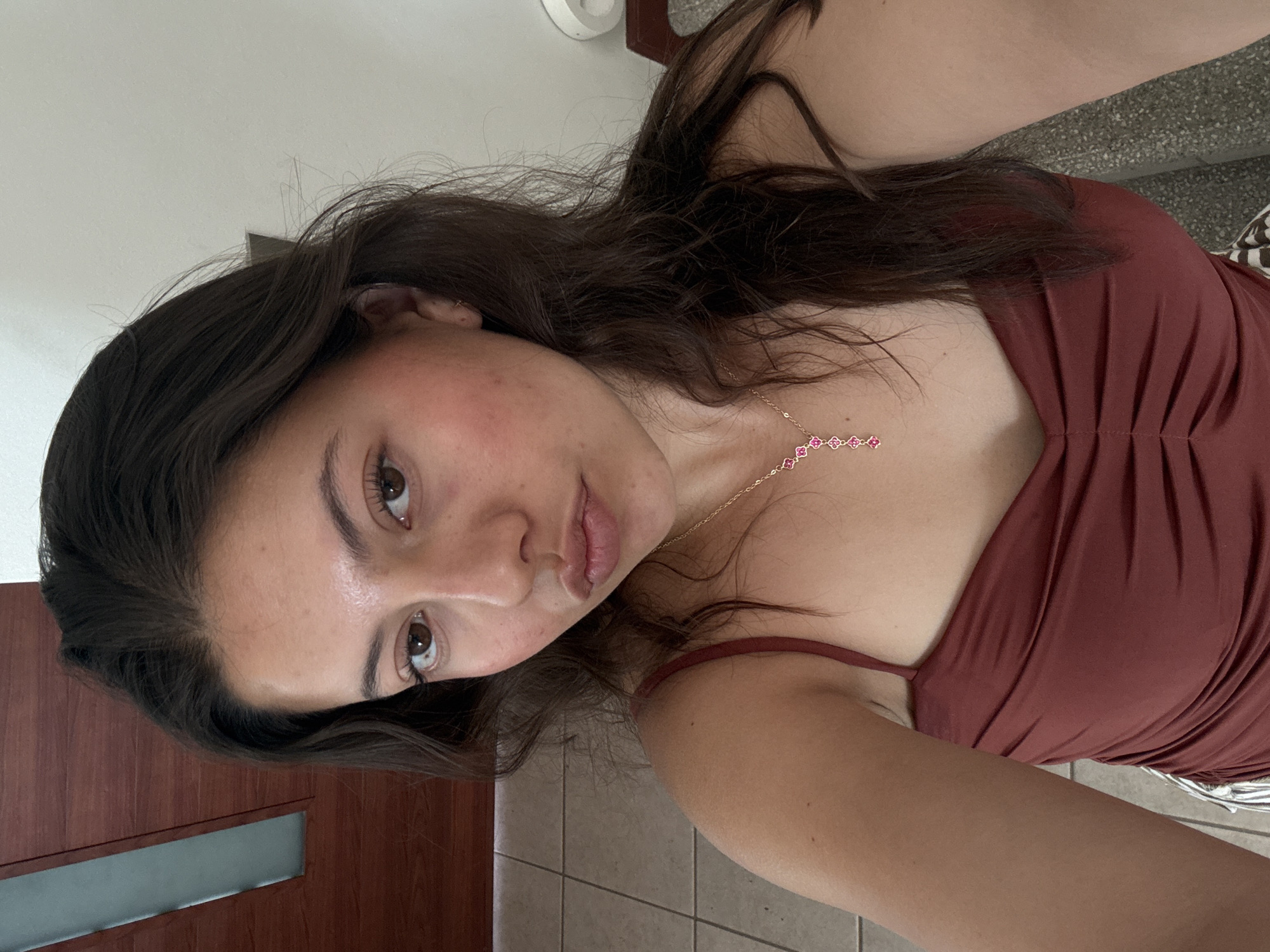 A young woman is taking a selfie. She has dark brown hair and is wearing a maroon-colored top. A gold necklace with pink flower-shaped details is visible around her neck. The background includes a door and tiled flooring. The location is unspecified and cannot be determined from this image.