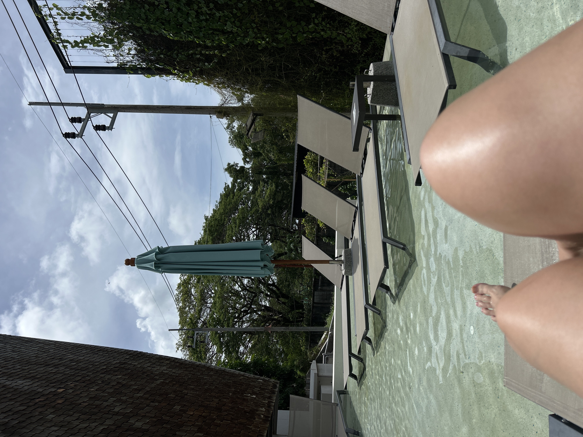 The image, likely taken in Rawai, Thailand, shows a partial view of an outdoor space with lounge chairs. The ground appears to be paved with a light green, water-like texture. A row of lounge chairs with light-colored cushions sits parallel to the frame, their black frames visible. An umbrella, light blue in color, is seen among the trees and a roof with brown shingles, partially visible in the lower left corner. Above the trees, utility poles and wires are present against a cloudy sky. A person's leg and foot are visible in the foreground.