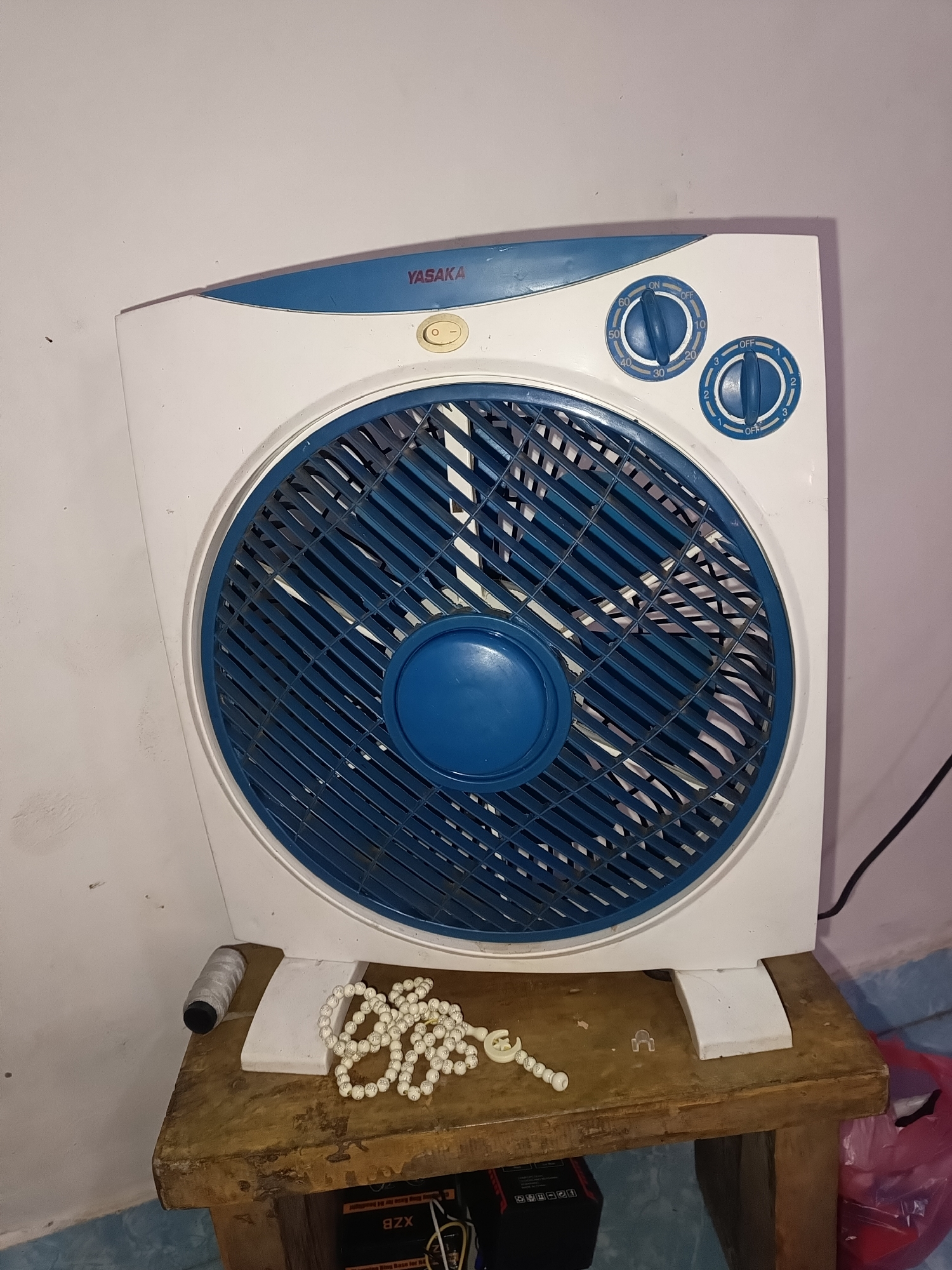An indoor, eye-level, medium shot captures a white and blue square-shaped electric fan positioned on a rustic wooden table. The fan features a blue grill and a circular blue central cap. Two blue dials are visible on the top right of the fan: one with settings marked from "OFF" to "60" and another from "OFF" to "3". A small yellow button is located centrally on the top of the fan, below the brand name "YASAKA". To the left of the fan's base, a spool of white thread with black ends rests on the table. A string of white beads, resembling prayer beads, is draped across the front of the table in front of the fan. The wooden table has visible signs of wear and tear. The background consists of a pale wall, appearing to be an interior setting, possibly in Terbanggi Marga, Indonesia, given the context provided. A black electrical cord extends from the fan, disappearing out of frame to the right. A pink plastic bag is partially visible on the floor to the far right.
