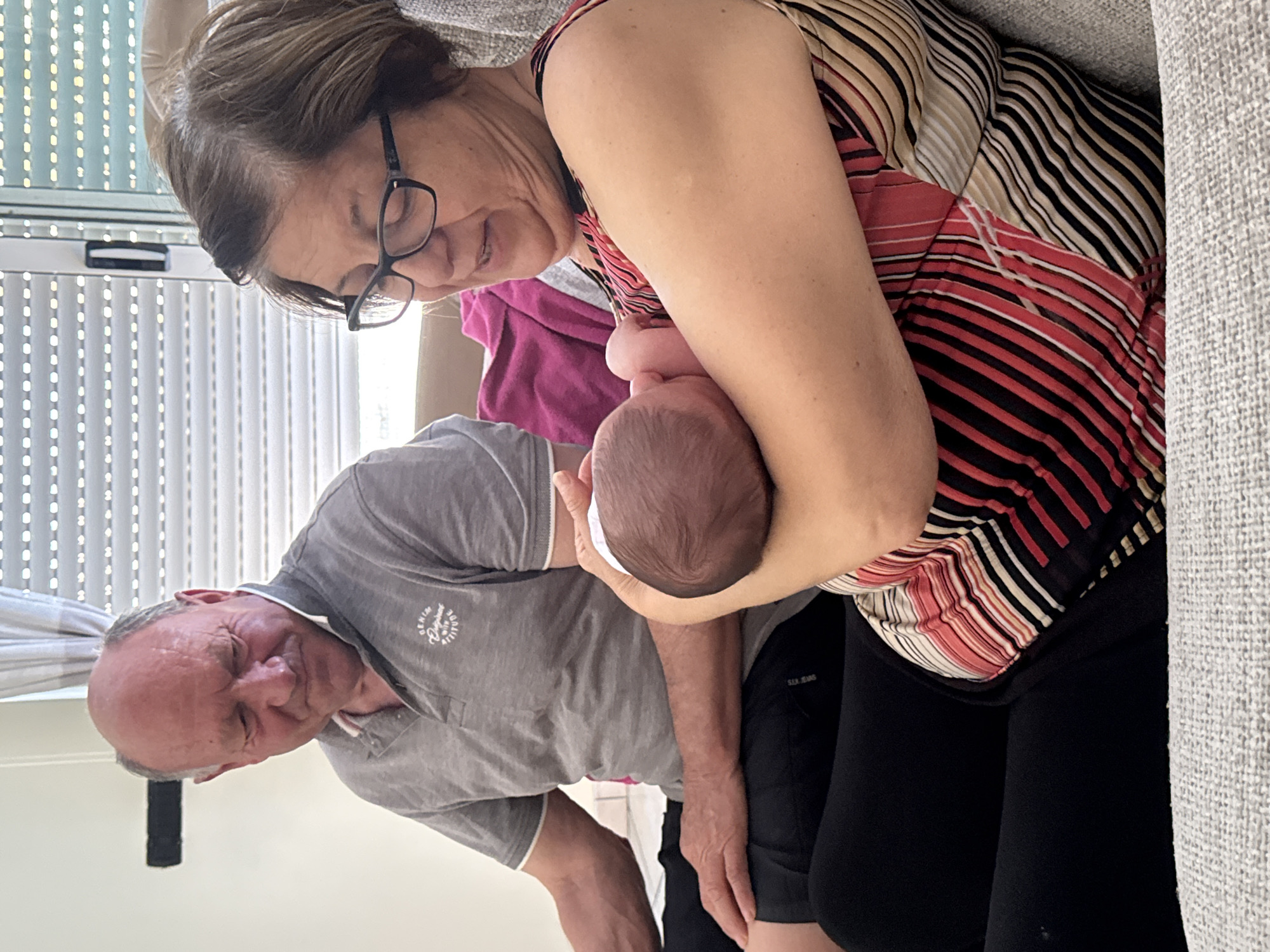 The image depicts two older adults, a man and a woman, holding a baby. The woman, wearing glasses and a striped shirt, sits on a couch with the baby in her arms. The man, in a grey polo shirt, leans in to look at the baby. The setting appears to be indoors, possibly a home in Aprilia, Italy. The background includes a window with blinds and a portion of the grey couch.