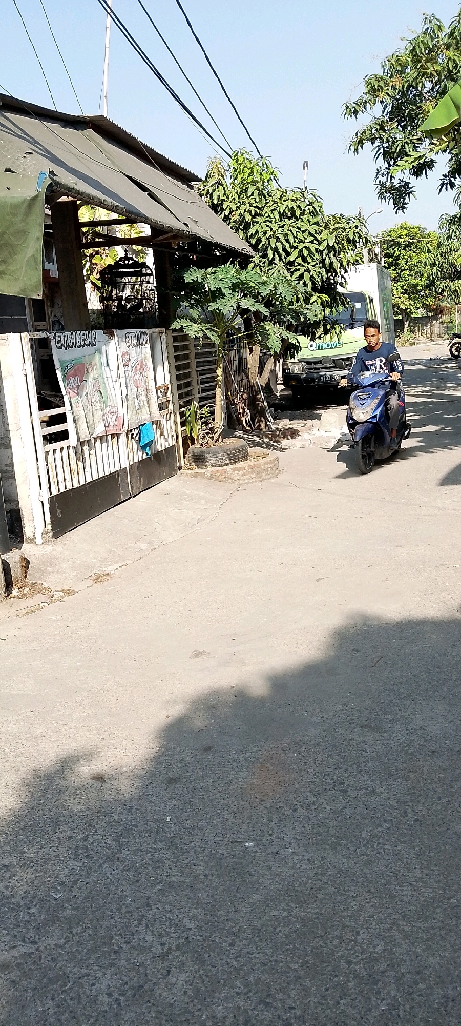 A young man rides a blue scooter down a paved road in Satriamekar, Indonesia. He is wearing a dark long-sleeved shirt and dark pants. The scooter has a license plate visible with the characters B 9703 BOB. To the left of the road is a white metal gate with black accents, behind which is a building with a dark, corrugated metal roof and several large posters affixed to the gate. To the right of the gate is a papaya tree planted in a tire. Further down the road, a white truck with the logo "Qmove" is parked. Trees with green foliage are visible on either side of the road and behind the truck. The sky is clear and blue. The scene appears to be a residential or commercial street.