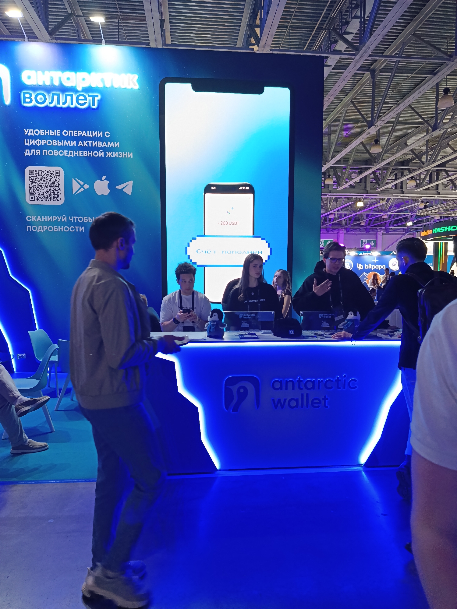 The image depicts a trade show booth for "antarctic wallet", located in Khimki, Russia. The booth is bathed in blue light and features a large backdrop with the company's logo and branding. A display shows a smartphone screen with a balance of -200 USDT. Several people are interacting at the booth, with staff members standing behind the counter and attendees approaching it. The environment appears to be a busy trade show floor with other booths and attendees visible in the background.