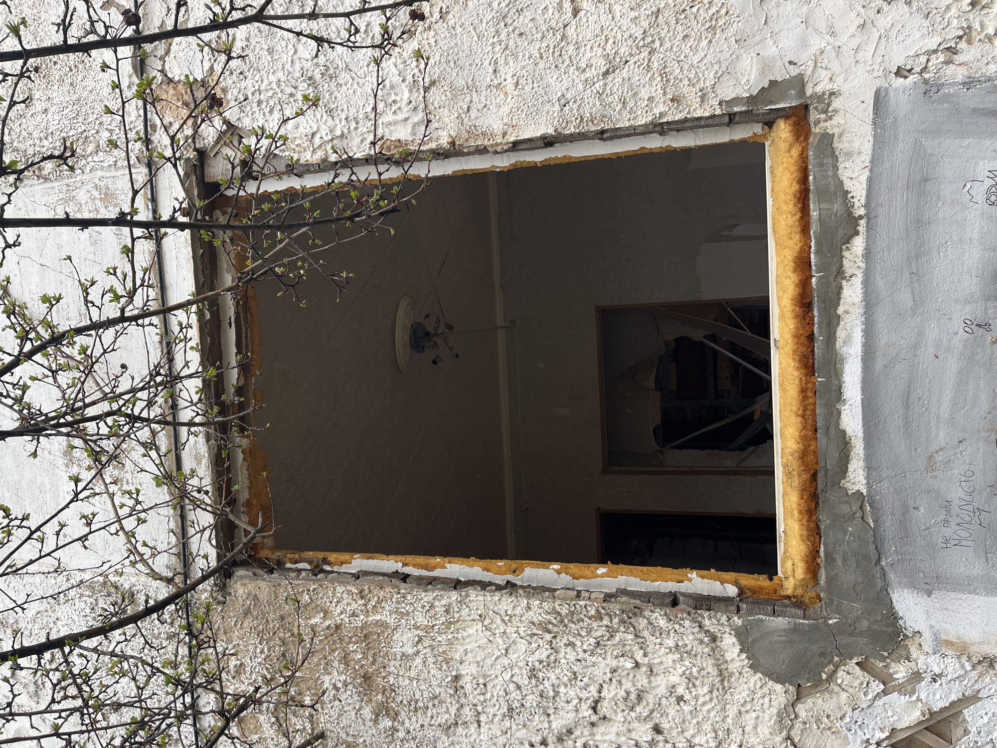 The image depicts a view through an empty window frame of a building in what is likely the city of Chaikovsky, Russia, as suggested by the Cyrillic inscription on the wall to the right of the window. The wall surrounding the window is rough and textured, covered in peeling white paint. The window frame is filled with yellow insulation. Inside, the room is dimly lit, revealing remnants of a chandelier on the ceiling and what appears to be debris and damage in the back room, visible through another window or doorway. A tree branch with budding leaves partially obscures the view on the left side of the window. The scene suggests abandonment or disrepair.