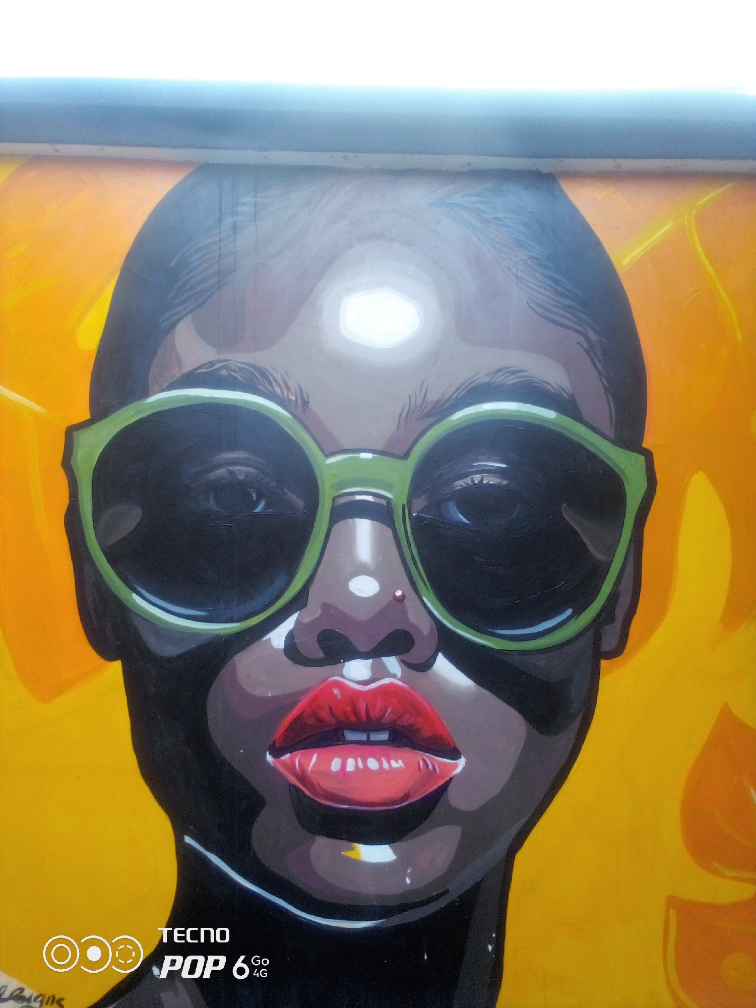 This is a close-up, waist-up, frontal portrait of a mural depicting a woman wearing large, lime-green sunglasses. The woman has dark skin, black hair, and striking red lips with a subtle shimmer. A small red stud is visible on her left nostril. The mural is rendered in a bold, graphic style with strong contrasts and defined lines. The background is a vibrant, abstract yellow and orange pattern that suggests organic shapes, possibly leaves or flowers. The mural is situated in Howayi, Nigeria. In the bottom left corner of the image, partially obscured by the mural's artwork, are white circular graphic elements and the words "TECNO POP 6 Go 4G" printed on what appears to be a wall or a signage. The lighting suggests it is daytime, with a bright, possibly overexposed sky visible at the top of the frame.