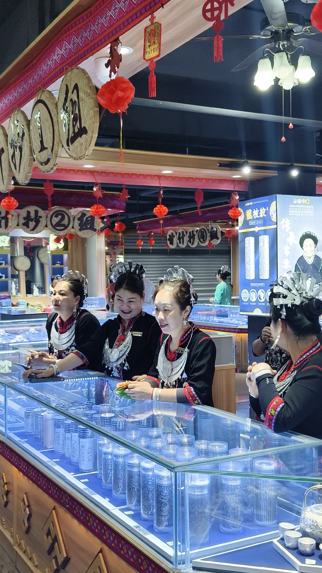 Inside a retail establishment located in Jiyang District, China, four adult females are positioned behind a long, illuminated glass display case. Each woman wears black traditional ethnic clothing adorned with large silver-colored necklaces and ornate silver-colored headwear. The second woman from the left exhibits a broad smile, while the third woman from the left also smiles and holds a small green and yellow item on the display case. The first woman from the left gazes to her right with a neutral expression. The woman on the far right is partially turned away from the camera, with her hands clasped.
The primary display case contains multiple clear cylindrical containers, each housing patterned silver-colored articles such as cups or small ingot-like pieces. A separate section of this case features a silver teapot and three small silver cups.
Overhead, red paper lanterns and red patterned banners with Chinese characters are suspended. Round, woven discs with prominent Chinese characters also hang from the ceiling structure. A multi-light ceiling fan is visible. In the background, a large blue and white promotional display board is visible, depicting traditional silver items and an elderly woman in ethnic attire. Chinese text on the board includes '龙族故' and '传统文化'. A fifth individual, dressed in a light green shirt, stands further within the retail space.