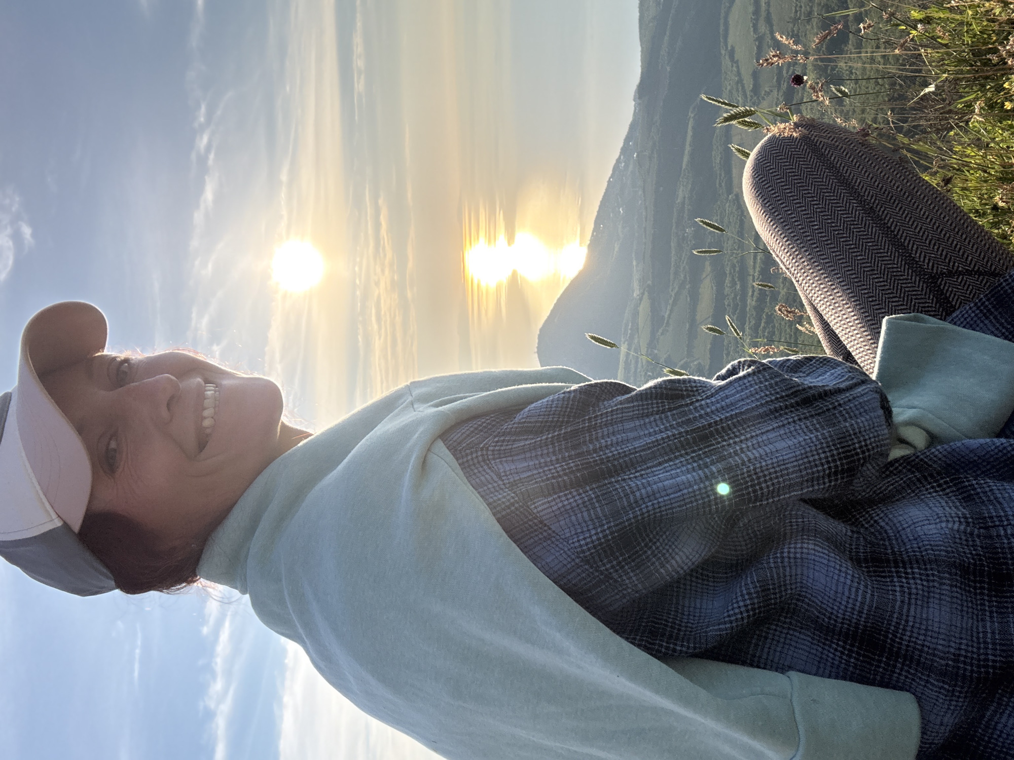 An outdoor portrait captured at either sunrise or sunset depicts a woman seated on a grassy hillside, looking over her left shoulder towards the viewer with a broad smile. The woman, with auburn hair visible beneath a two-toned baseball cap (white/grey and light pink/beige), is illuminated by the golden light of the low sun positioned in the upper right quadrant of the image. She wears a light blue or mint green sweatshirt draped over her shoulders, covering a blue and grey plaid button-up shirt. Her legs are bent, revealing patterned pants, likely a herringbone or chevron weave, in shades of brown and beige.
The background features a vast body of water, possibly an ocean or large lake, reflecting the bright sun in a distinct vertical streak of light. Mountains or hills rise between the observer's position and the water, showing some sparse vegetation and faint signs of human development. The immediate foreground consists of various grasses and wildflowers, with some tall, seed-headed stalks and a few small purple flowers visible. The sky is clear blue with scattered wispy clouds. A small green lens flare is present on the woman's plaid shirt. The scene is located in Unknown, Unknown.
