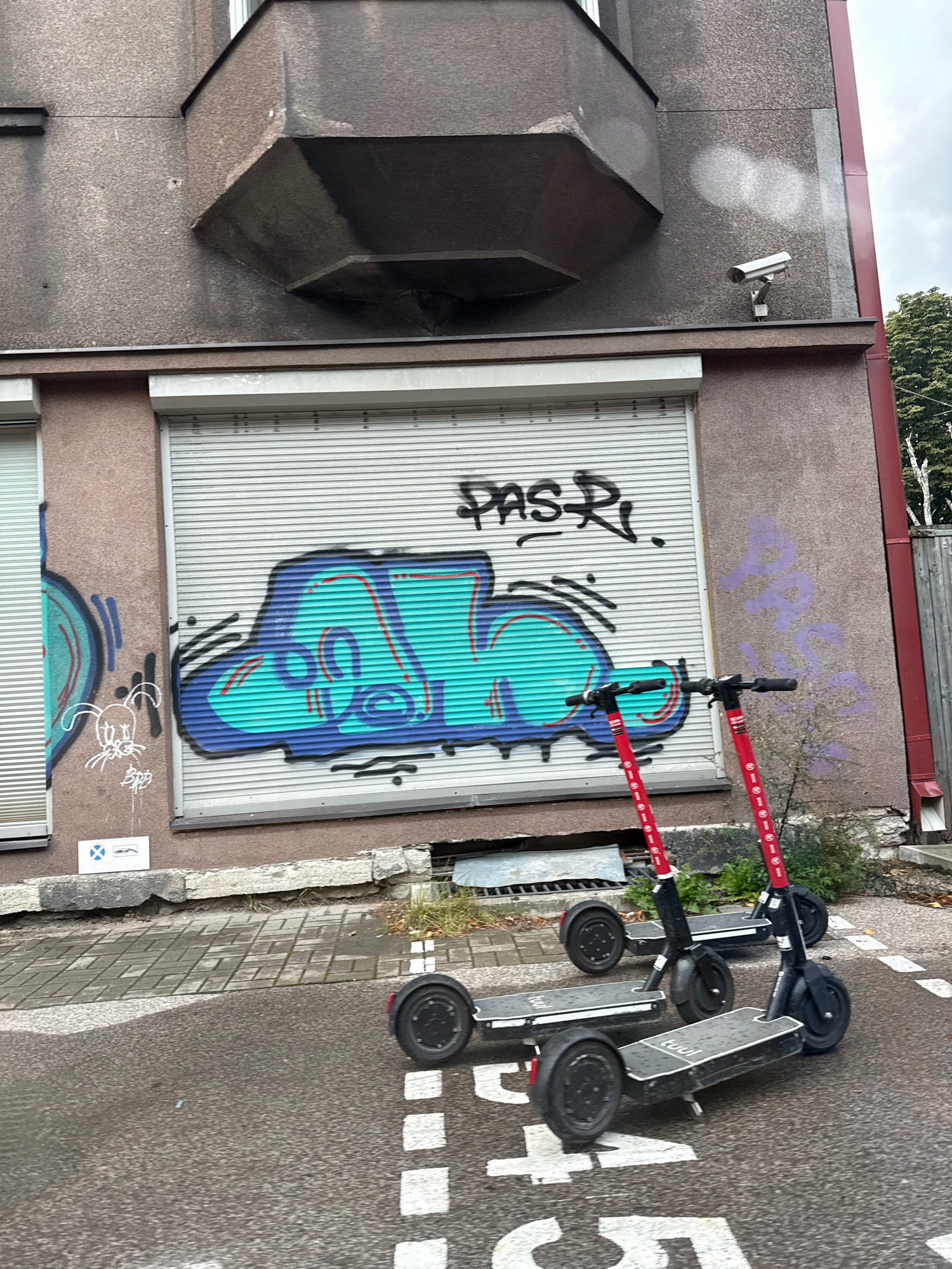 Two electric scooters are parked on a wet asphalt surface in front of a building with a large, graffiti-covered metal garage door. The scooters are red and black. The building's facade is textured concrete and has a small, gray security camera mounted on the upper right portion. The garage door has graffiti in teal and blue with red accents, as well as black lettering that reads "PAS R.". To the left of the garage door is a window covered by a white blind, and further left is another window with a blue and teal graffiti tag. A small blue and white sign is posted on the wall below the window. The ground in front of the garage door is paved with rectangular stones. The sky is overcast. The scene is located in the Kalamaja district of Tallinn, Estonia.