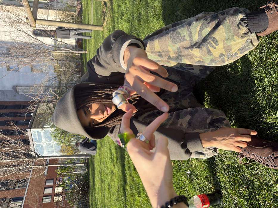 A person with dark skin and dreadlocks, wearing a dark olive green hooded sweatshirt and camouflage pants, sits cross-legged on a sunlit green lawn. They look directly at the camera with a slight smile and open mouth. Their right hand is extended forward, with the index finger and thumb forming a half-heart gesture. This hand displays a large silver-colored ring on the index finger and multiple rings with red stones on the middle finger. A second person's left hand, adorned with a large green ring on the index finger, a silver ring on the middle finger, and a black beaded bracelet, extends from the left frame, completing the heart gesture with the central figure's hand. A red Coca-Cola bottle is visible on the grass near this second hand. The central figure wears dark brown laced shoes.
In the background, a park-like setting features bare trees, indicative of late autumn or winter. To the left, a modern brick building with windows is visible. A glass structure, possibly a greenhouse or modern architectural feature, stands in the mid-ground. To the right, a significant historic stone building with arched windows, characteristic of older Parisian architecture, is present. Two other individuals are visible in the background: one seated on the grass to the left, and another standing further back on the right. The scene is illuminated by bright sunlight, casting distinct shadows. The location is identified as the Jardin Anne-Frank in the 3rd arrondissement of Paris, France.