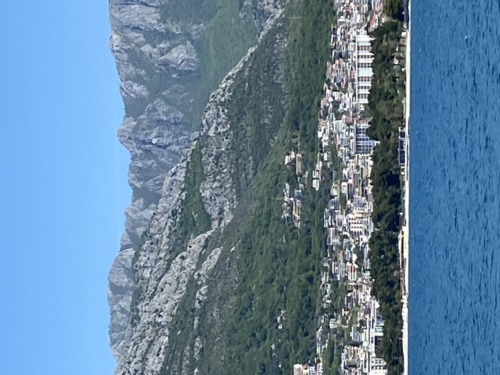 This image depicts a vibrant coastal landscape, oriented as a panoramic view. A clear, light blue sky occupies the top portion of the frame. Below this, a majestic, rugged grey mountain range with sharp, rocky peaks stretches across the scene. The mountain slopes transition into dense green vegetation, which then gives way to a sprawling urban settlement.
The town, nestled between the mountains and the sea, features numerous buildings of varying sizes and architectural styles, predominantly light-colored. These structures are densely packed, indicating a significant coastal development, likely comprising residential buildings and possibly hotels.
A narrow, light-colored strip of land, appearing to be a beach or promenade, separates the town from the sea. The water is a deep blue, exhibiting gentle ripples across its surface. The scene is brightly illuminated, suggesting it is daytime with clear, sunny weather. There are no visible people or discernible activities taking place, and no text is present in the image. The combination of steep mountains, a built-up coastline, and the sea is characteristic of the Adriatic region, aligning with the location context of Bar, Montenegro.