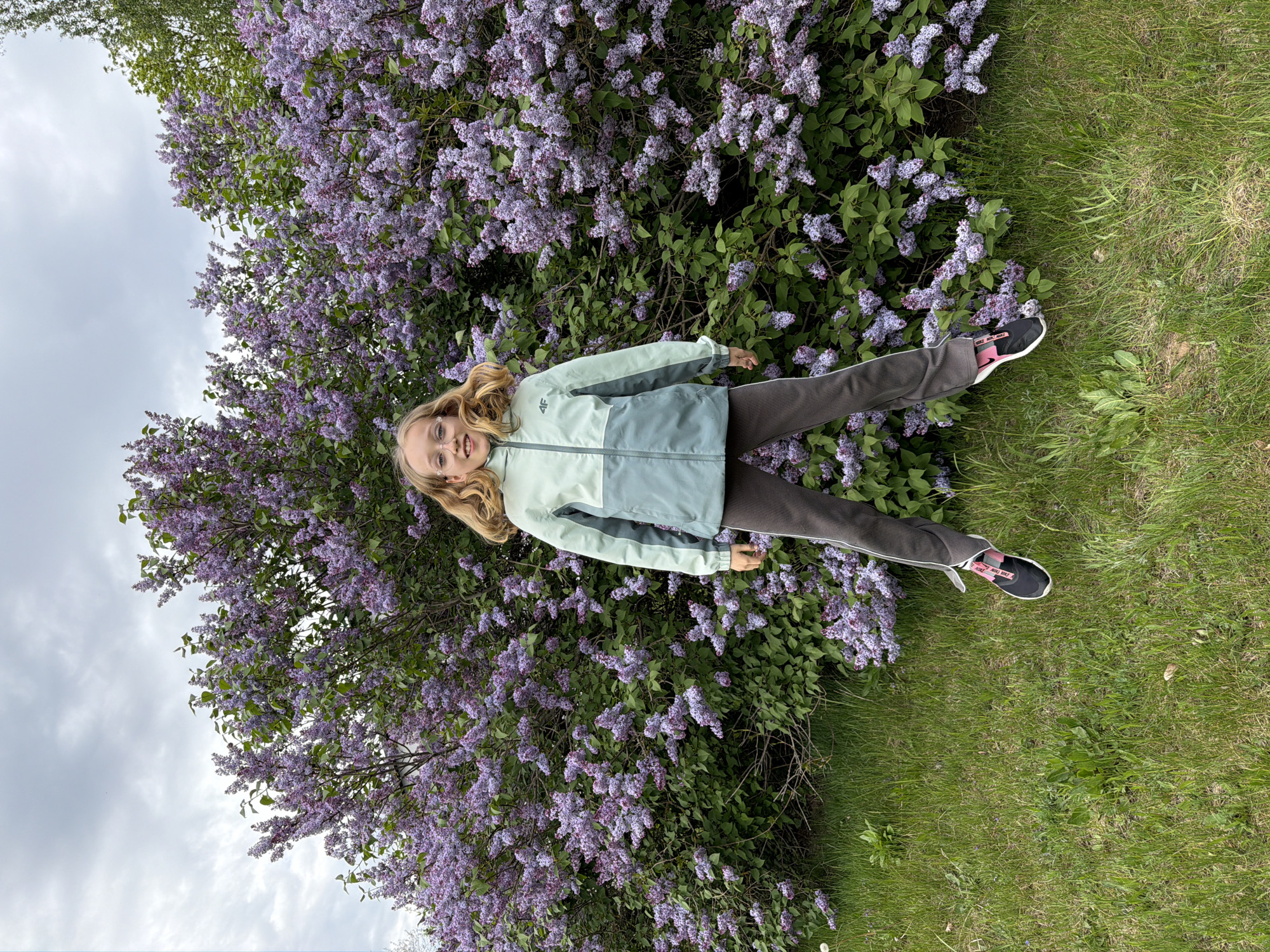 A young girl lies in front of a blooming lilac bush in Dreiliņi, Latvia. She wears glasses, a light green and gray jacket, dark pants with a white stripe, and black and pink sneakers. The lilac bush is in full bloom with purple flowers. Green grass surrounds the bush. The sky is cloudy.