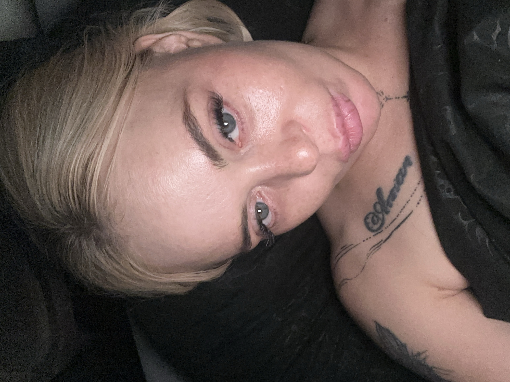A woman with blonde hair pulled back and makeup, including false eyelashes and lip gloss, is shown in a close-up, low-angle selfie. She has a tattoo on her left shoulder and collarbone, and another tattoo on her neck. The background is dark and appears to be indoors, possibly a bed. The lighting is dim, with a strong light source coming from above and to the right, creating highlights on her face.