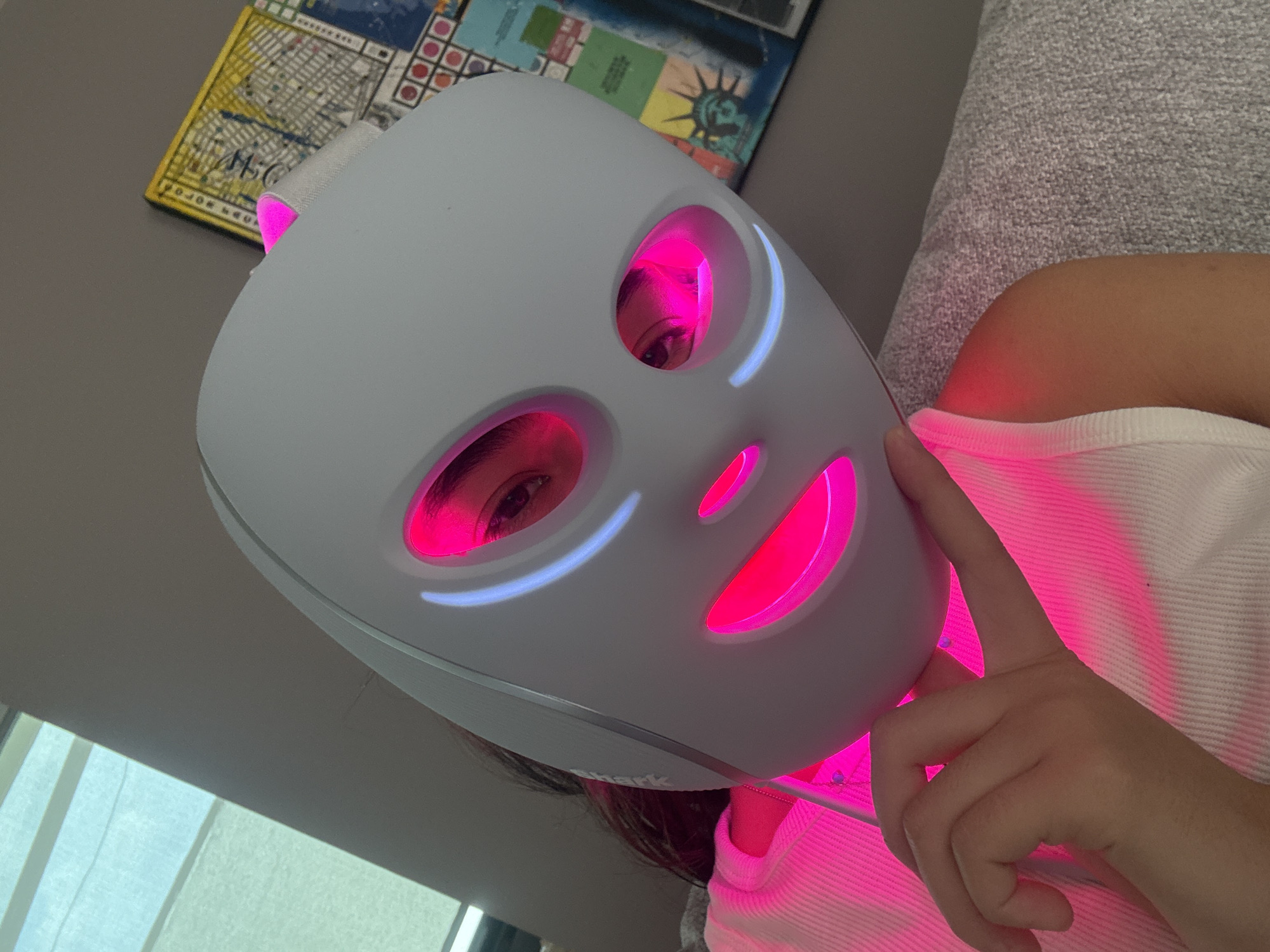 A person is wearing a light therapy mask with red and blue lights. The mask covers the majority of their face. The person is lying on a gray textured surface. They are wearing a white tank top. A hand is visible near the mask. Behind the person, there is wall art featuring a map and the Statue of Liberty. The location is not San Bernardino Tlaxcalancingo, Mexico.