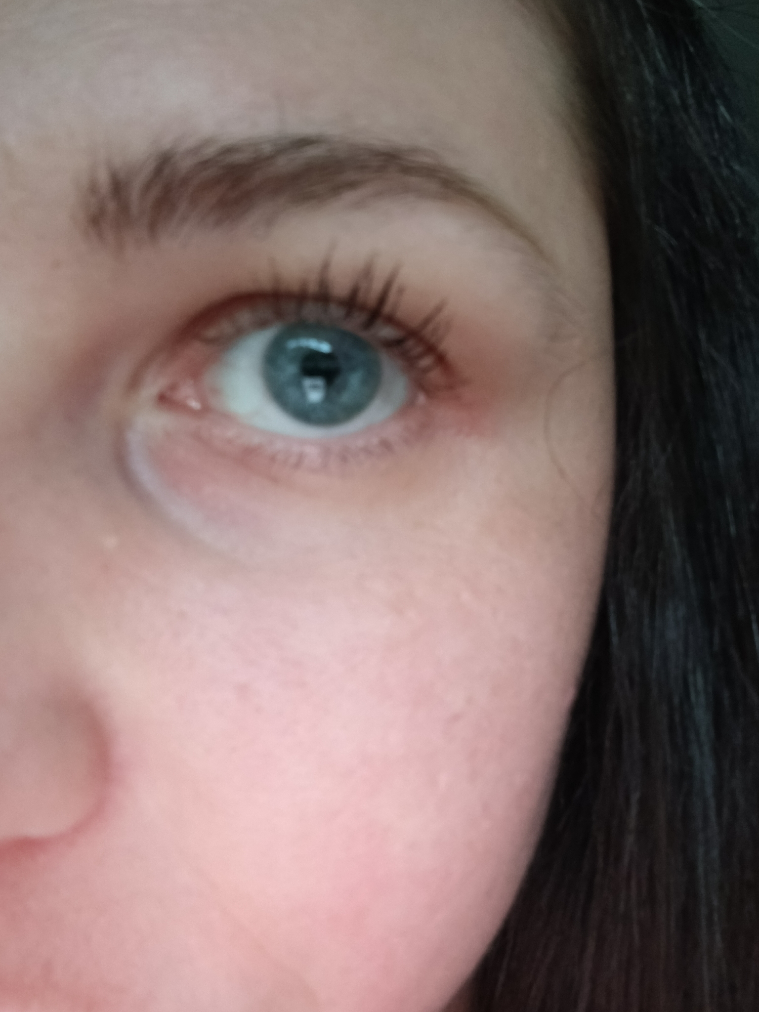 A close-up of the right side of a person's face, focusing on the eye. The eye is a blue color with dark lashes. The eyebrow is visible above the eye and is brown in color. The skin is fair. Dark hair is visible on the right side of the frame. The location is Unknown, Unknown.