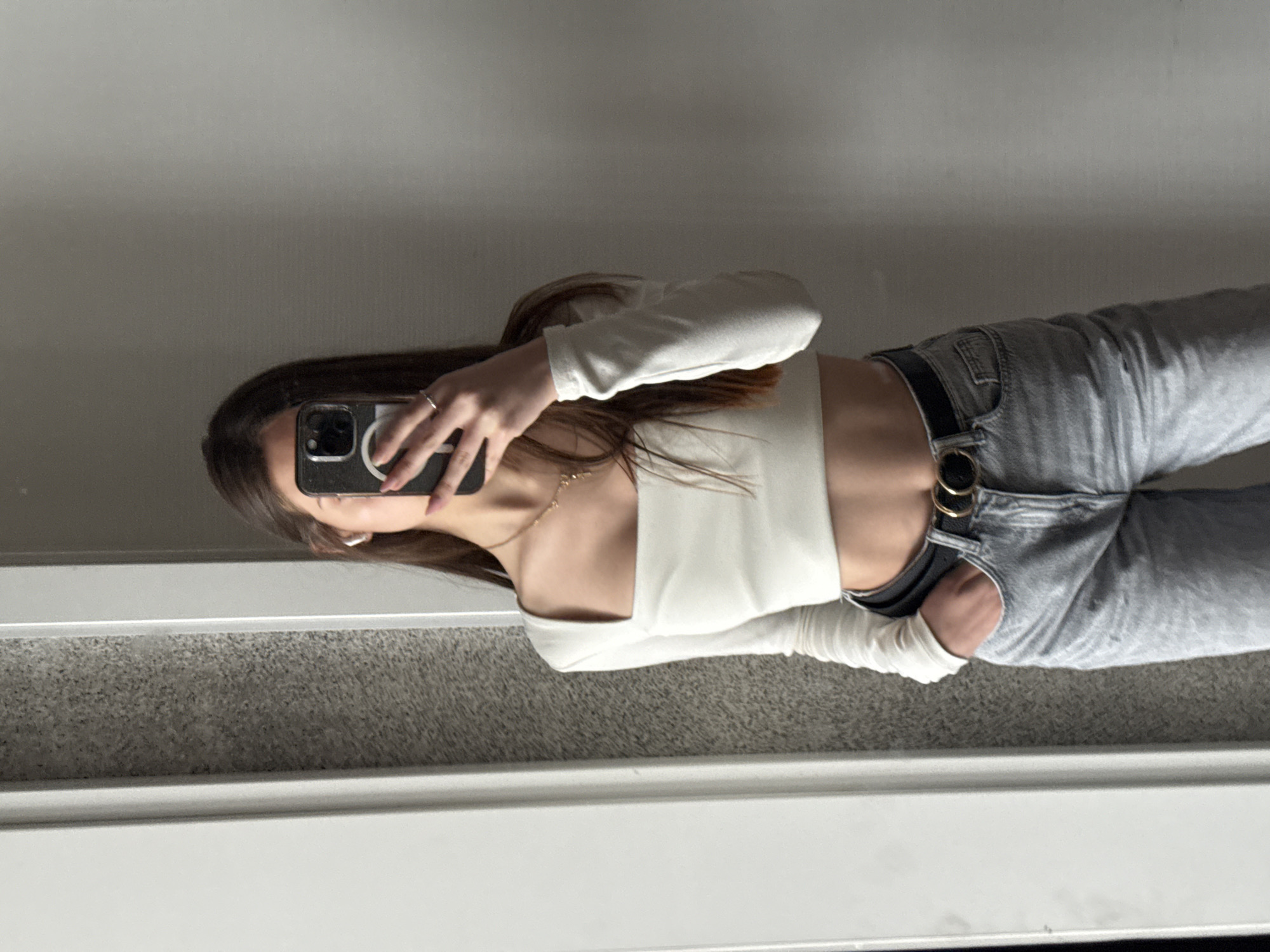 A young woman stands in front of a mirror and takes a photo with her smartphone. She is wearing a white long-sleeved crop top and grey jeans with a black belt. The background appears to be a room with a grey floor. The city of Sofia, Bulgaria cannot be determined from the image.