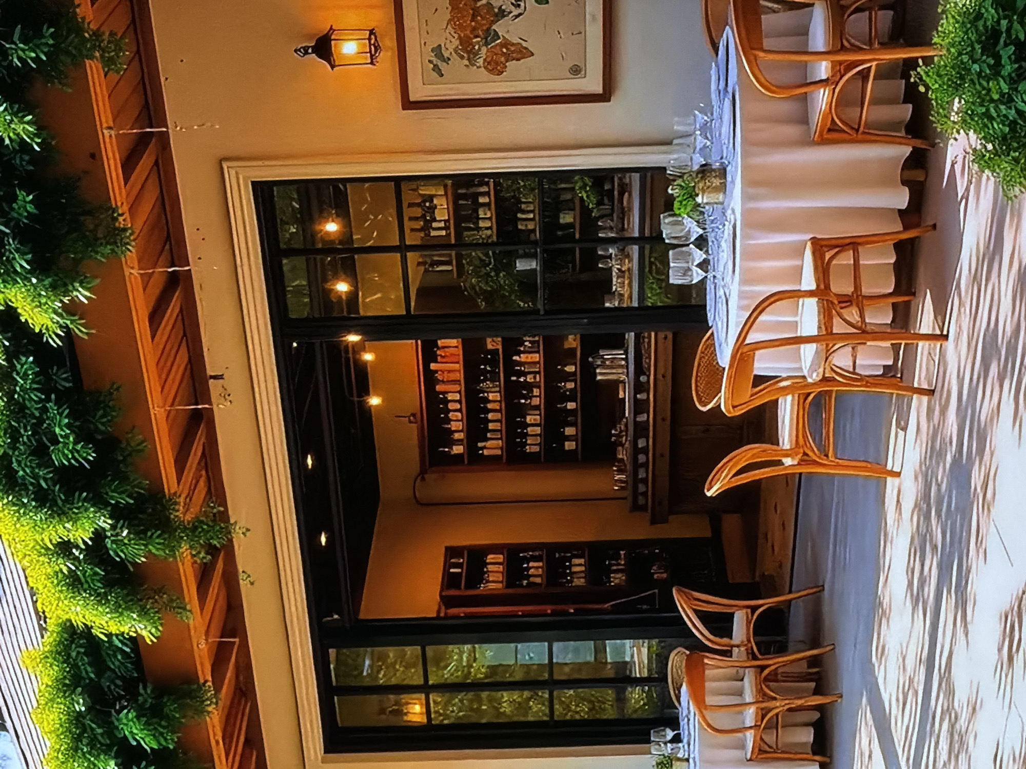 An inviting outdoor patio area of a restaurant or cafe is depicted in Sofiivska Borshchahivka, Ukraine. The scene features at least two round dining tables, elegantly covered with white tablecloths, set for guests. Each table is adorned with multiple wine glasses and a small green centerpiece. Light-colored wooden chairs with curved designs are arranged around the tables.
The patio is situated next to a building with a cream-colored facade, from which lush green foliage cascades downwards along wooden support structures. A decorative, warm-glowing lantern-style wall sconce is mounted high on the exterior wall. Above the large, dark-framed glass windows and doors, a framed artwork, possibly a map or an abstract piece with earthy tones, is visible. Through the clear glass, the interior reveals shelves heavily stocked with wine bottles, indicating a well-supplied wine collection or cellar within the establishment.
The ground of the patio is a paved or concrete surface, displaying distinct shadows, suggesting natural light, possibly during late afternoon or early evening. The overall ambiance is elegant and serene, prepared for a dining experience. No people are visible in the image, and there is no discernible text.