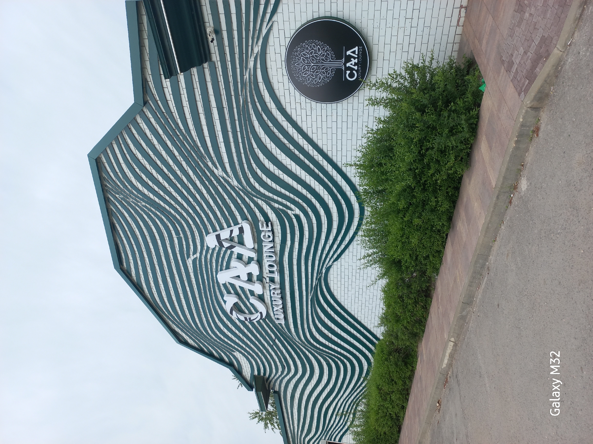 The image shows the exterior of "CAFÉ LUXURY LOUNGE" in Volzhsky, Russia. The building has a modern design with a facade featuring wavy, horizontal lines in white and teal. A round sign with a tree motif and the letters "C&A LUXURY LOUNGE" is mounted on the white brick wall. Green bushes grow along the base of the building. The sidewalk and part of the road are visible. The photo was taken with a Galaxy M32 smartphone, as indicated in the lower right corner.