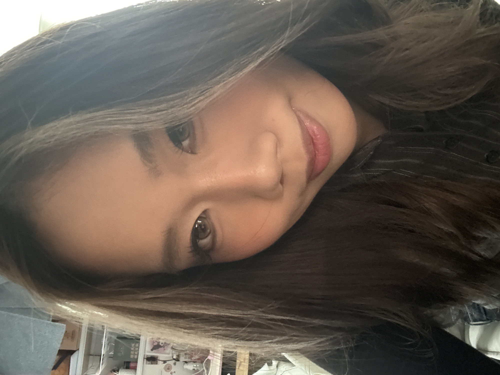 The image is a close-up selfie of a young woman with long, dark brown hair that is slightly parted. Her head is tilted, with her eyes looking directly at the camera. She has fair skin and is wearing a subtle lip color and blush. She is wearing a dark shirt with a subtle pattern. The background is blurry, showing glimpses of a cluttered desk with boxes, various containers, and other items. The location, Nan'an District, China, is not discernible from the image.