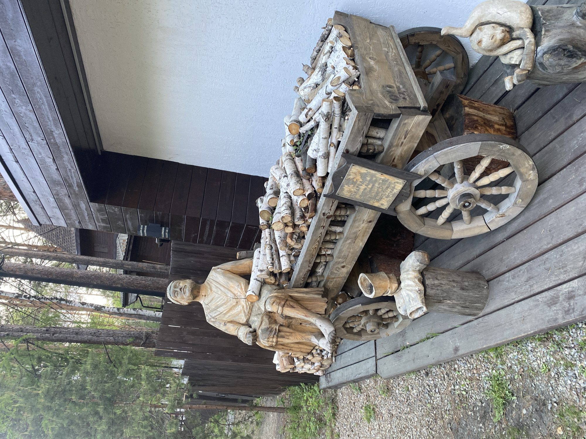 The image depicts an outdoor scene in Tal'tsy, Russia, featuring a wooden sculpture of a man sitting on a cart filled with logs. The cart has wooden wheels and is made of rough-hewn planks. The man sculpture is carved with detailed clothing and facial features, and a few logs are placed beside him as if he is holding them. Other wooden carvings, including a cat sculpture, are visible on the wooden platform where the cart is situated. The background includes a wooden fence, tall trees, and the exterior of a building with dark wooden siding and a white plastered section. The ground is covered with gravel and some vegetation. The scene appears to be part of an outdoor exhibition or decorative display.