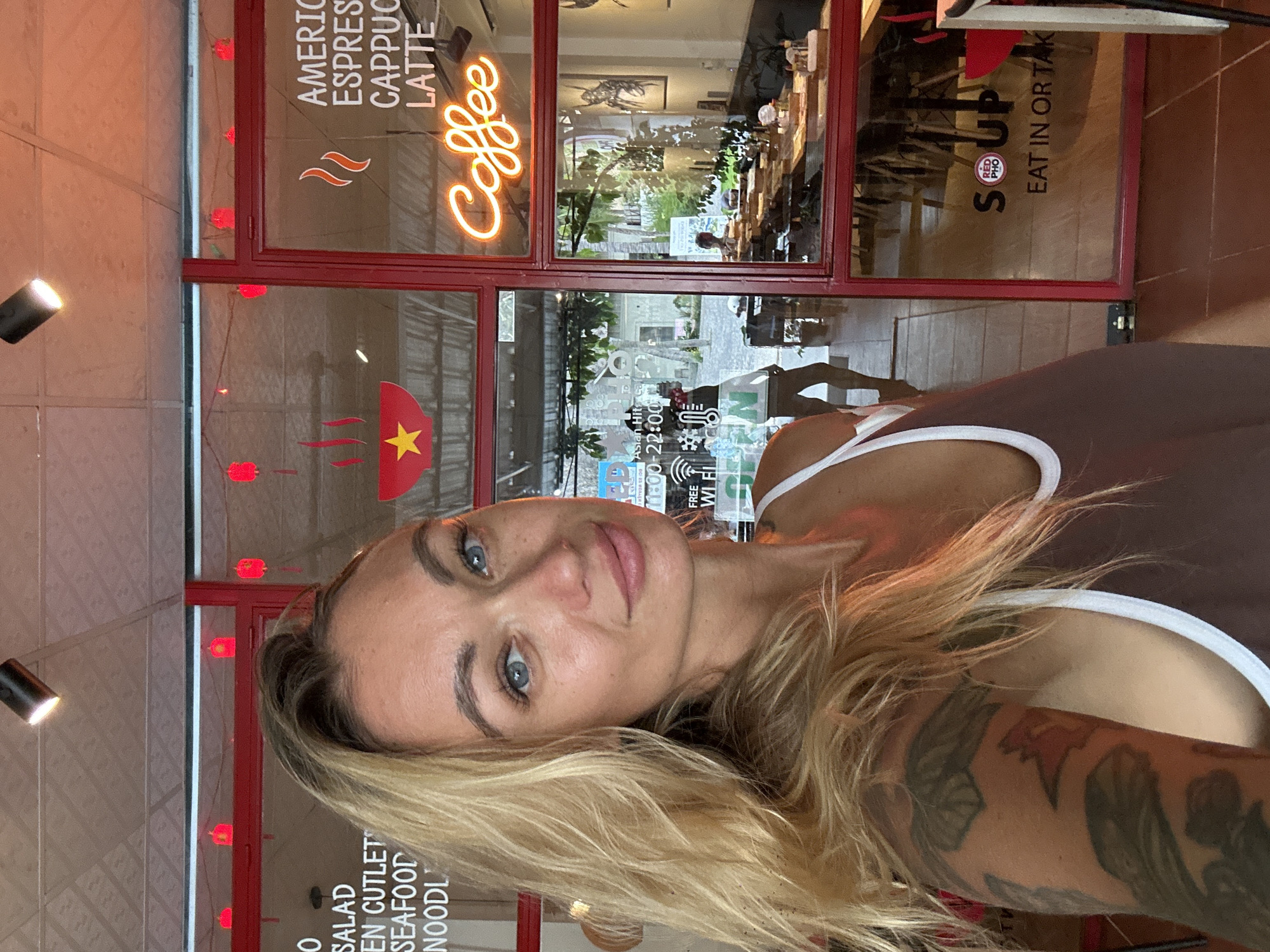 A woman with blonde hair and blue eyes takes a selfie in front of a restaurant in Phu Thuy Ward, Vietnam. She is wearing a brown tank top. The restaurant has red frames around its windows and doors. Text on the window reads "SALAD", "SEAFOOD", "NOODLE", "AMERICAN ESPRESSO", "CAPPUCCINO", "LATTE", and "Coffee". A sign also says "SOUP" and "EAT IN OR TAK". Inside the restaurant, tables are set with food and chairs. Several people can be seen through the windows. Red lanterns hang above the window.