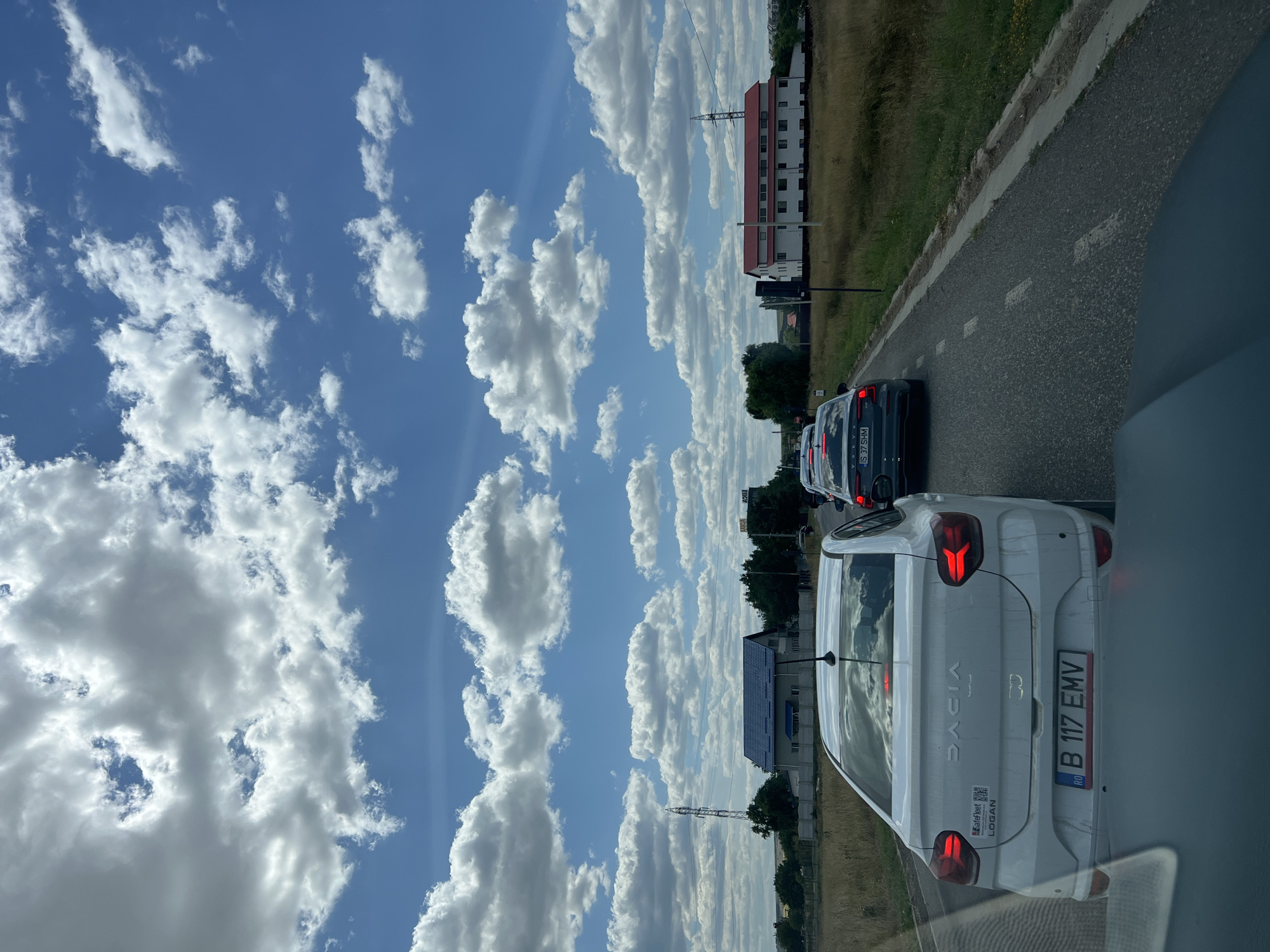 The image depicts a traffic jam on a two-lane asphalt road in Podu Iloaiei, Romania. The sky is filled with white, puffy cumulus clouds against a bright blue background. In the foreground, a white Dacia Logan is stopped, with its brake lights illuminated. The license plate reads "B 117 EMV". Behind it, a line of vehicles, including a dark-colored Volvo, are also stationary.
The surrounding environment consists of dry, grassy fields on either side of the road, with scattered trees and bushes. In the distance, several buildings are visible. To the left, there is a house with a blue roof and a tall transmission tower. To the right, a large, white building with a red roof stands out. The road has visible white dashed lane markings. The overall impression is a hot, sunny day with a stalled traffic situation.