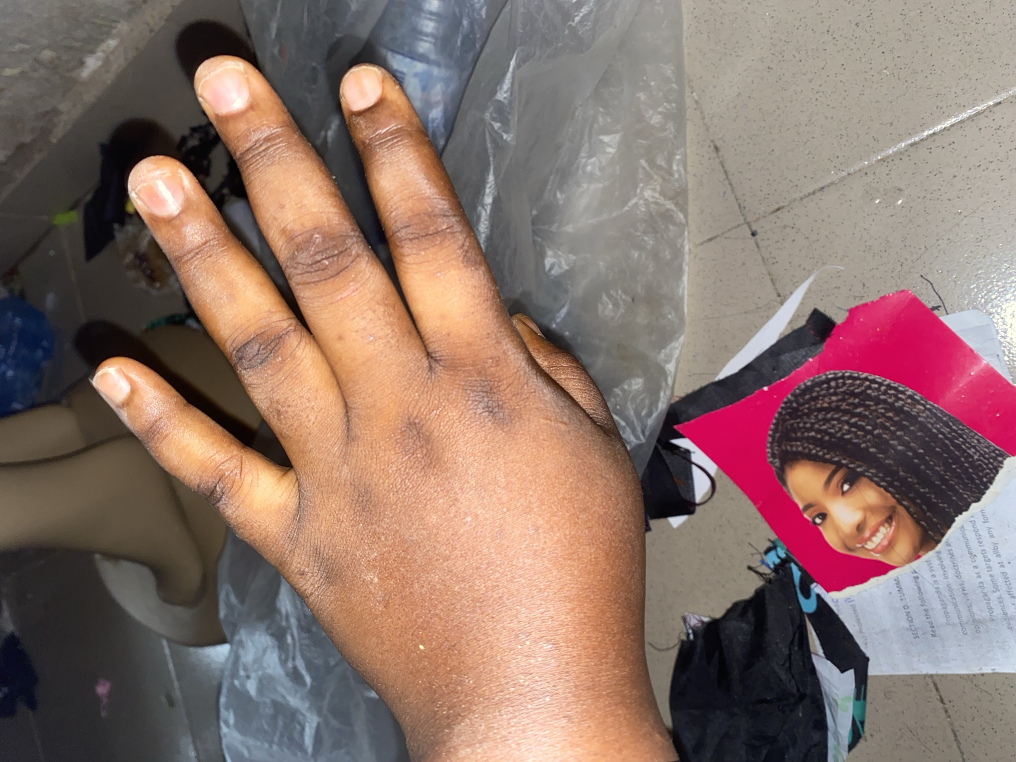 A dark-skinned hand, with four fingers extended and the thumb tucked, is positioned in the foreground. The hand exhibits visible skin texture and some dryness, particularly around the knuckles and finger joints. The fingernails appear unpolished and natural. The hand is resting on a surface that transitions from a pale, slightly speckled floor to a crumpled, translucent plastic sheeting. In the background, out of focus, is a beige, mannequin-like form, possibly a torso or leg, and various discarded items including black fabric and a blue plastic bottle. To the bottom right of the hand, a ripped piece of pink paper with an image of a smiling woman with braided hair is visible, partially obscuring some text from a document below. The scene appears to be an indoor setting, with the floor suggesting a potentially commercial or public space. There is no indication of the specific location in Jauro Yinu, Nigeria, as the image lacks any discernible landmarks or context for that precise locale.