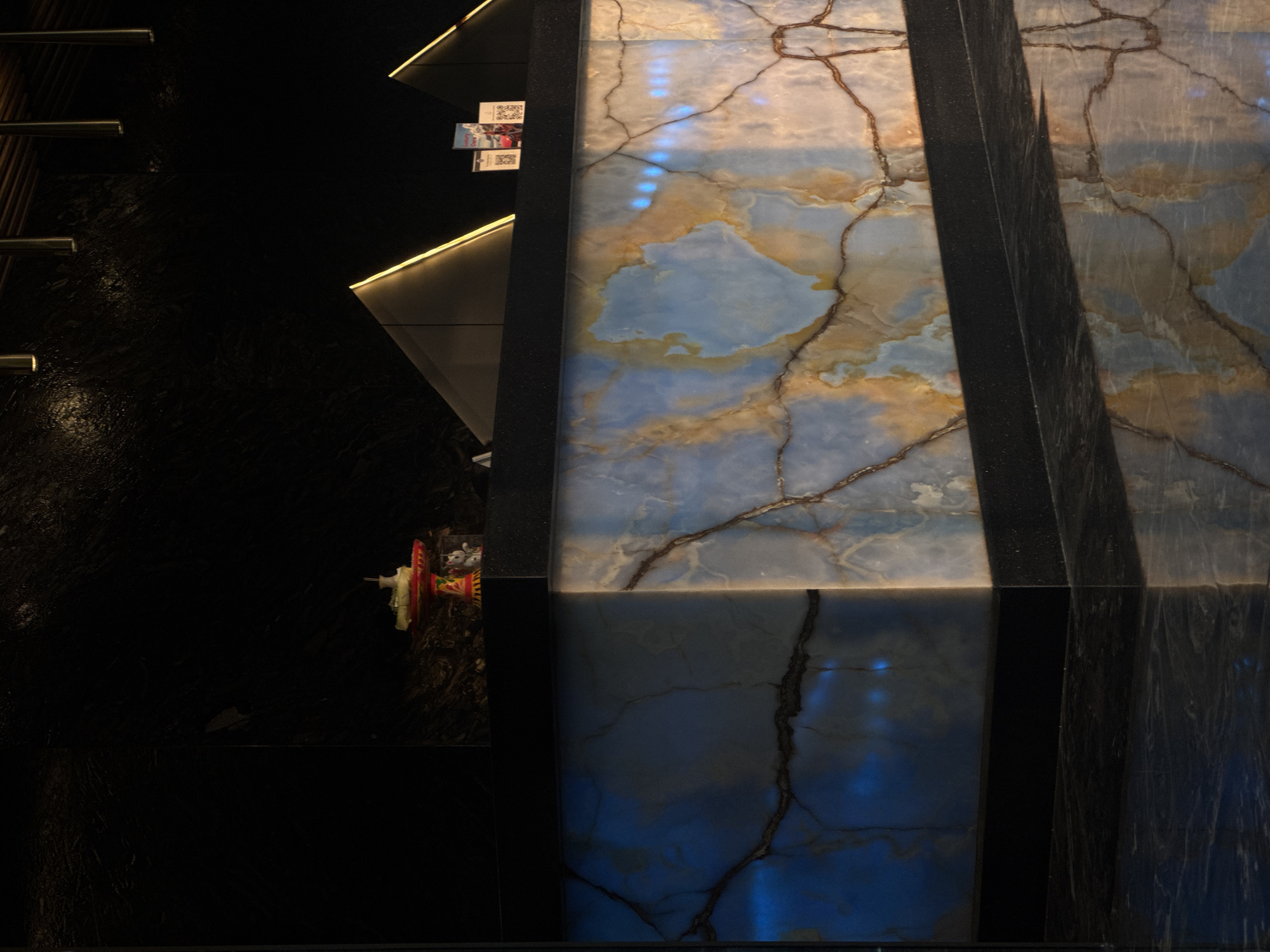 An interior architectural scene showcases a modern design featuring backlit natural stone panels, likely onyx or marble, with prominent blue, white, and golden-brown hues interwoven with dark, intricate veining. These radiant panels are vertically oriented and bordered by dark, textured surfaces, one of which incorporates a thin strip of warm yellow LED lighting, creating a sharp, illuminated edge.
To the left of the stone panels, large sections of highly polished black material, possibly dark granite or lacquered wood, reflect subtle light. Further left, several slender, horizontal metallic bars or railings are visible.
On an angled, dark shelf or ledge immediately adjacent to the backlit stone, a few small printed items are displayed. One of these clearly features QR codes, and another has the word "Luxury" partially visible, suggesting promotional material or informational brochures.
Near the bottom center of the frame, against the dark backdrop, a small, colorful decorative object or figurine is partially discernible. It appears to be crafted from red and yellow elements, topped with a small white figure, possibly an animal or character.
The image captures a static, luxurious setting, possibly a hotel lobby or high-end commercial space in North Kuta, Indonesia. The strong artificial lighting, particularly the backlighting of the stone, suggests an indoor environment where the time of day is irrelevant to the ambiance. The materials and sleek design convey a sense of modern elegance and sophistication. No people are present in the scene.