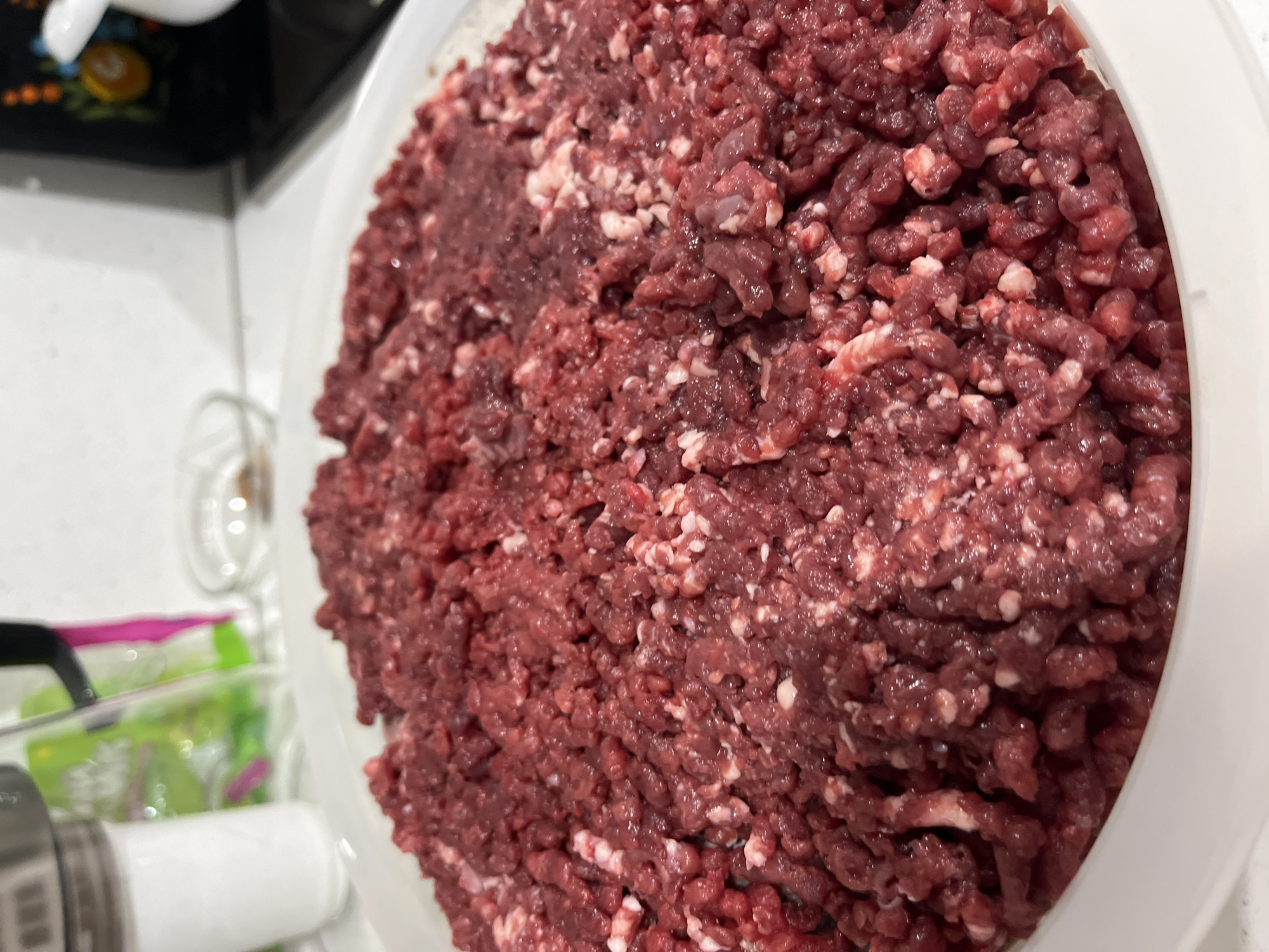 The image shows a large bowl filled with ground beef. The meat is a dark red color with flecks of white fat. The bowl is white. The background is blurry and shows part of a kitchen counter with other objects including a seasoning shaker, a container of salad, a transparent measuring cup, a black object with floral designs and a white counter top. There is no evidence of a location in Chaikovsky, Russia, or any people.