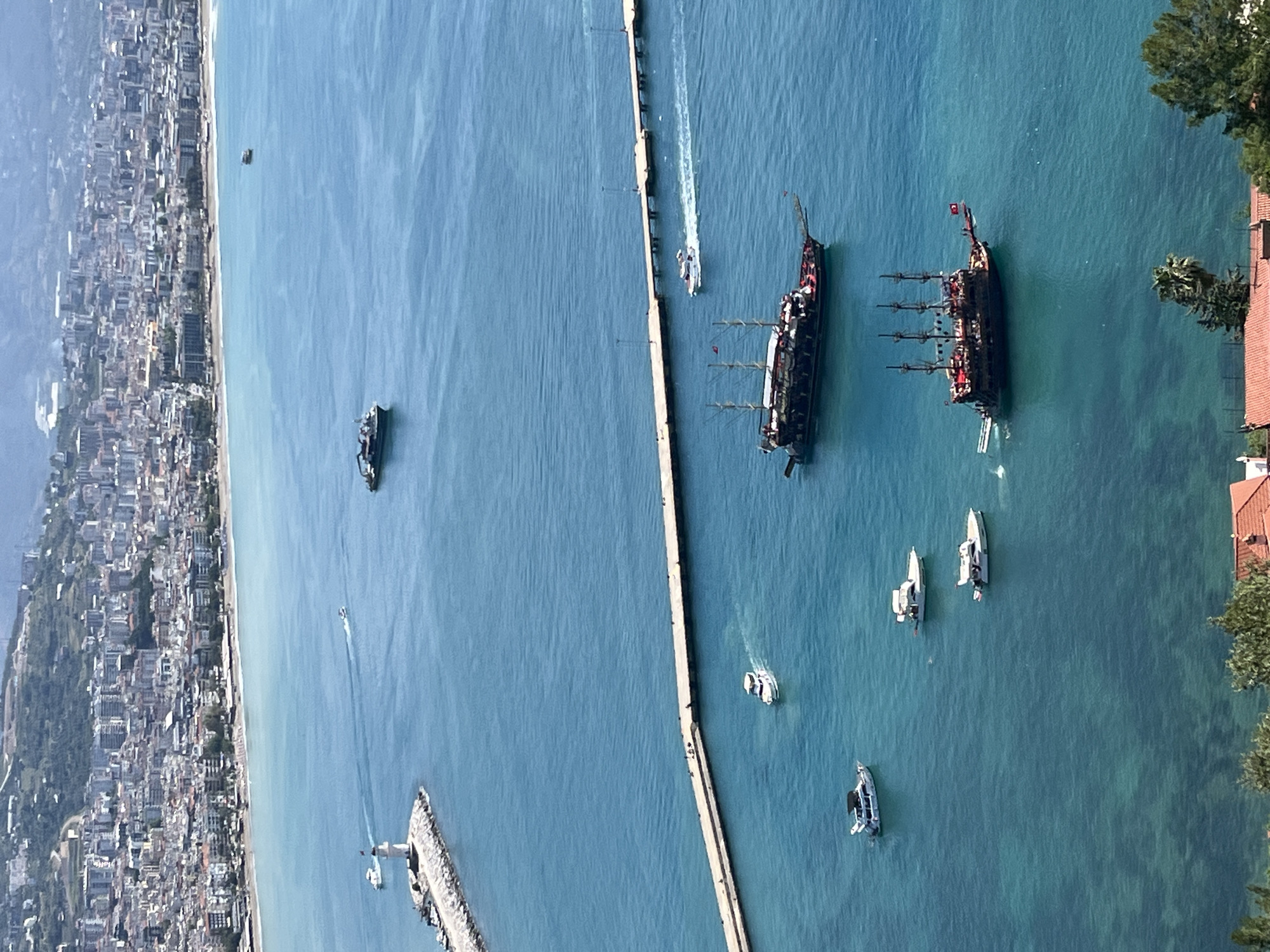 The image shows an aerial view of the coastline and harbor area of Alanya, Turkey. A long pier extends into the turquoise sea, separating the open water from the harbor. Along the coastline to the left, a dense urban landscape is visible, comprised of closely packed buildings and interspersed with greenery. Several boats are scattered in the sea. Two large pirate-themed ships are anchored near the coastline, suggesting a tourist attraction. Smaller boats and yachts are also present, indicating recreational or commercial maritime activity. The overall scene suggests a vibrant coastal city with a mix of tourism, urban development, and maritime activities. Vegetation lines the eastern side of the harbor.