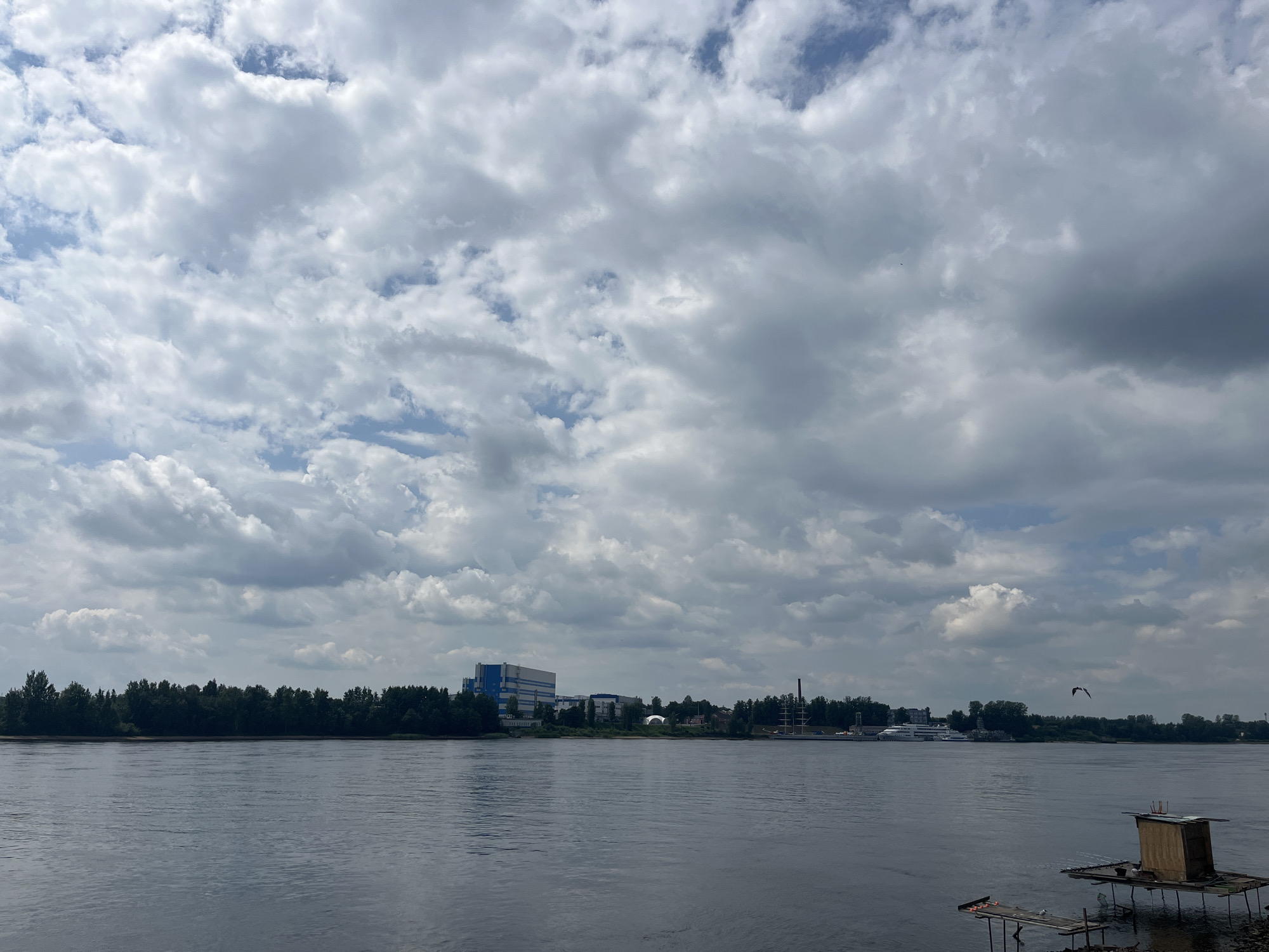 The image shows a wide river under a partly cloudy sky. On the far bank, a dense line of trees is visible, with a few industrial or commercial buildings peeking through in the distance. Among these buildings, a prominent structure is a large, modern blue and white building. Further along the riverbank, a tall smokestack and a large white yacht or ship are also visible. In the foreground, on the right side of the river, a small wooden structure on a raft is floating on the water, with another smaller platform with fishing rods nearby. A bird is captured mid-flight in the sky. The overall scene suggests an industrial or semi-industrial waterfront area. This scene is consistent with the location in the city of Imeni Sverdlova, Russia, which is situated along the Neva River and has a mix of industrial and natural landscapes.
