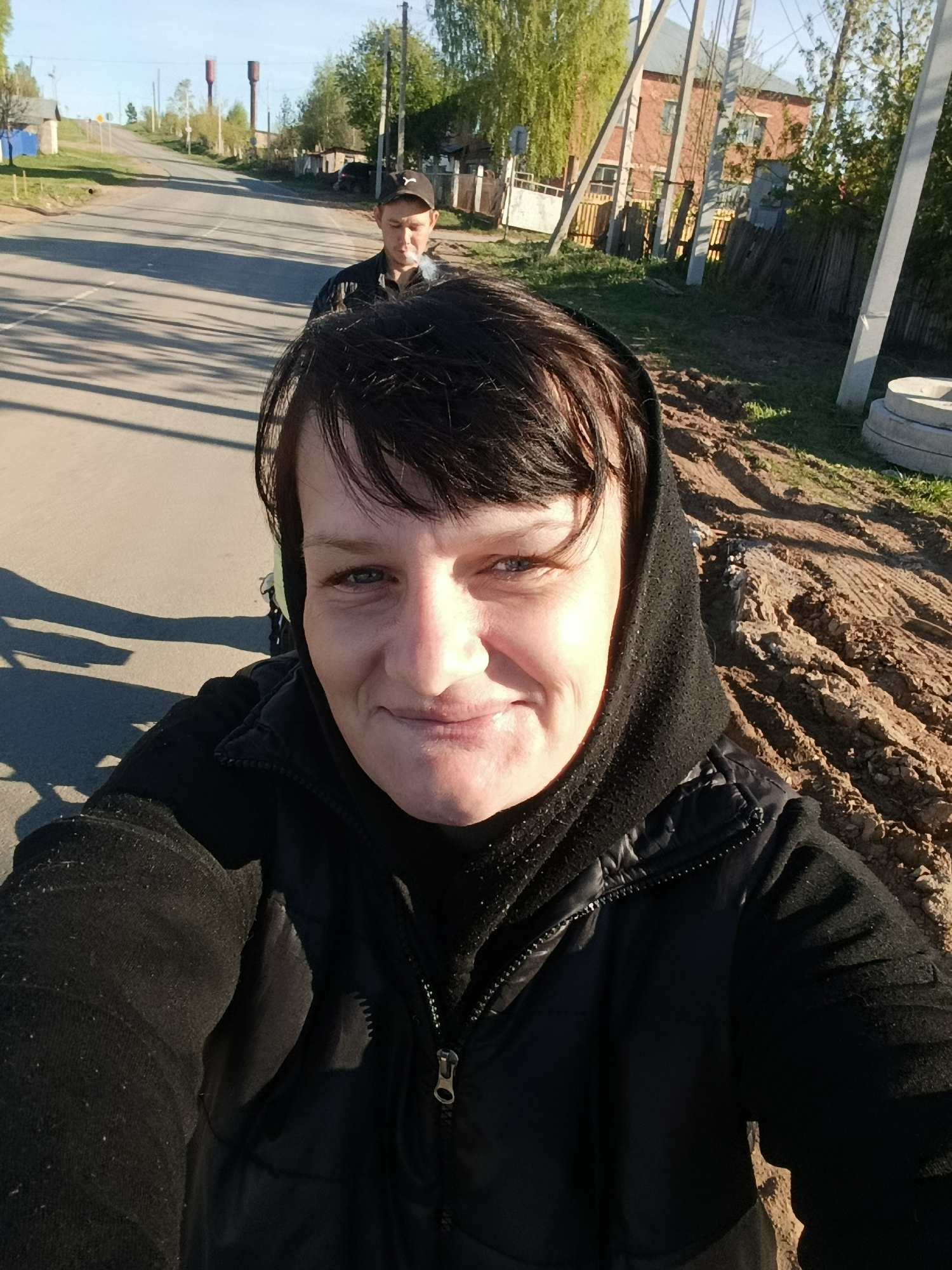 A selfie shows a woman wearing a black hooded garment and zip-up vest, positioned on the side of a road in Фоки, Russia. The road, stretching into the distance, shows another individual with a baseball cap. The background includes a mix of foliage, utility poles, and some buildings on the periphery. To the right, the terrain appears to be a dirt area.