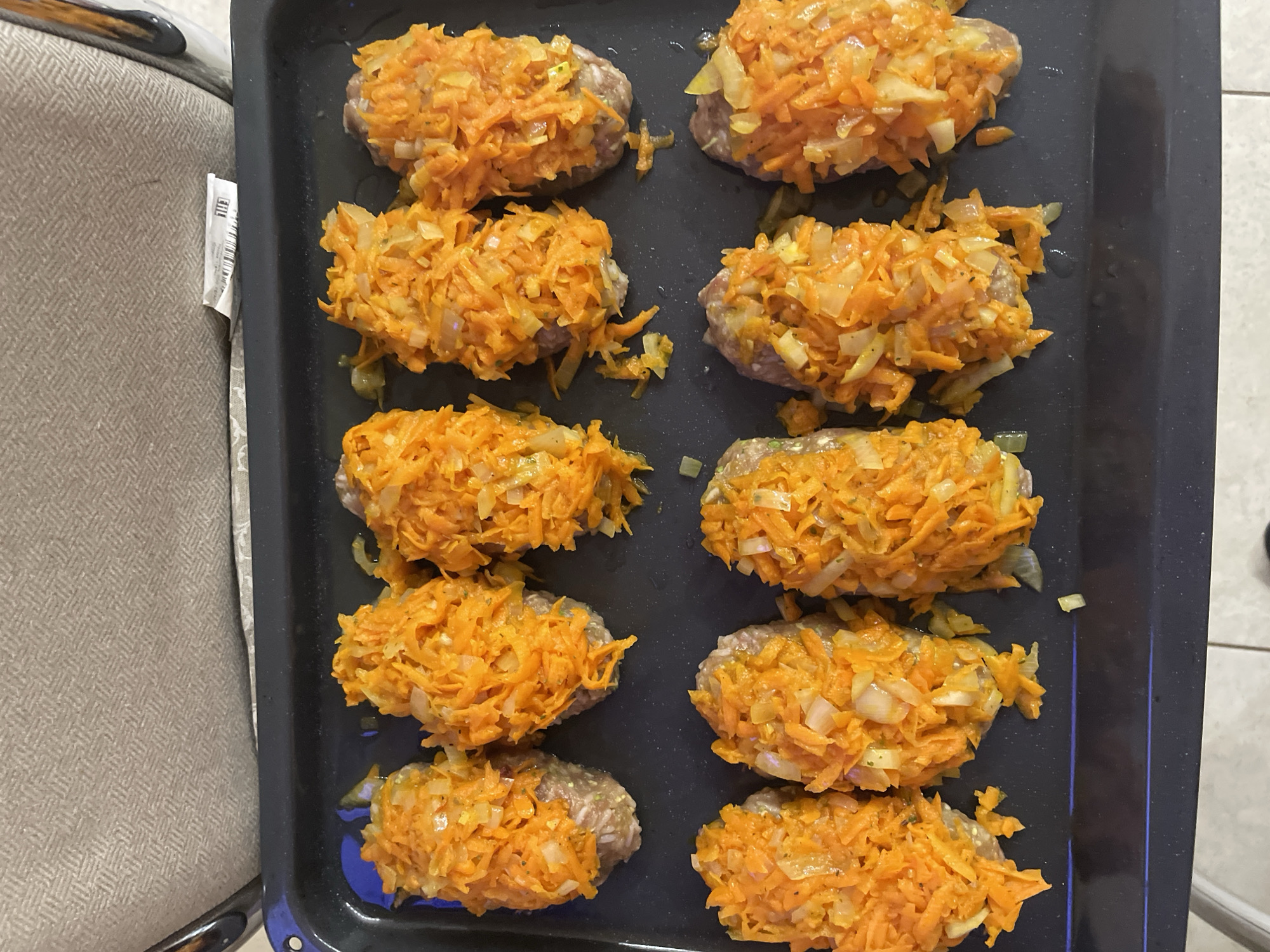 The image shows a black baking sheet containing ten meat patties topped with shredded carrots and onions. The baking sheet is resting on what appears to be a fabric surface next to a chair. The location can not be confirmed with the given information, but it is claimed to be Moskovskiy, Russia.