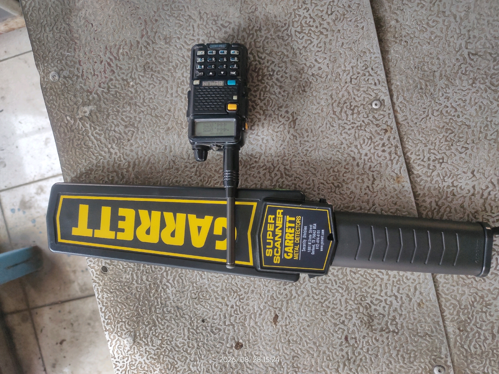 A handheld metal detector, a Garrett Super Scanner, is laid flat on a textured surface, possibly a metal sheet or mat. A small, black portable radio with a digital display and keypad is mounted vertically on top of the metal detector. The radio appears to be powered on, displaying numerical data on its screen. The overall scene is indoors, with what appears to be a tiled floor visible in the background on the left side of the image. The setting does not provide specific visual cues to determine the location in the city of Telagaasih, Indonesia.