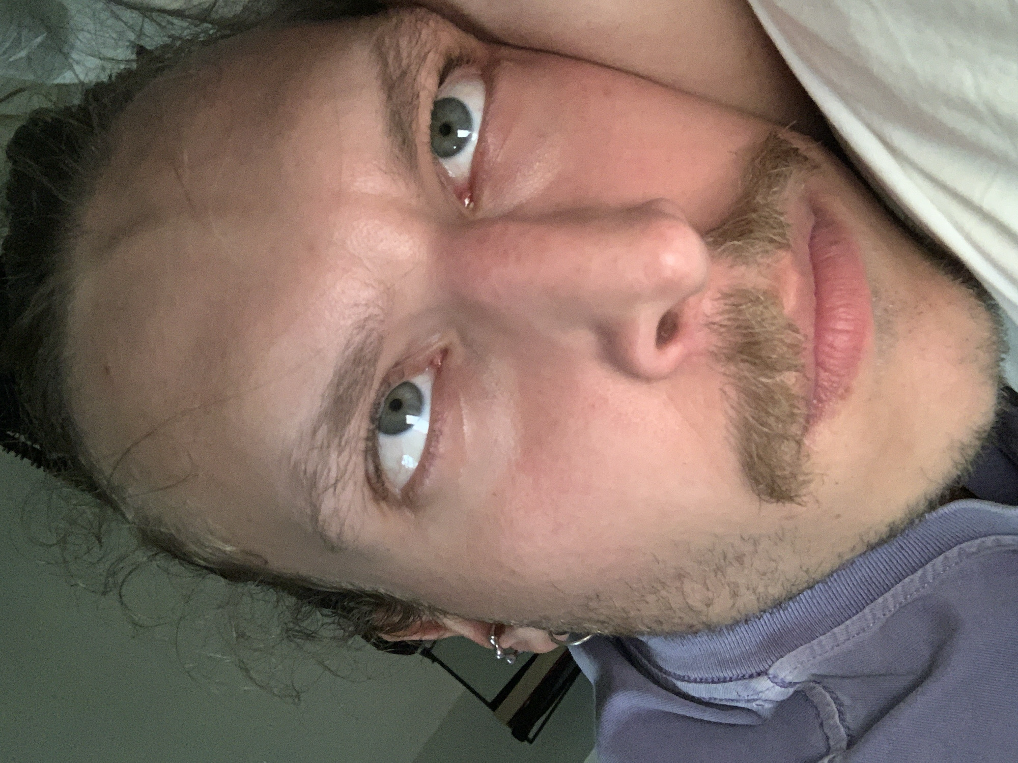 A close-up, indoor portrait of a fair-skinned male with light brown hair, a mustache, and light-colored eyes. He appears to be lying down, as indicated by the angle and the presence of a white object (possibly a pillow or bedding) in the upper right corner. He is wearing a blue or purple collared shirt. There is no visible indication that this is Tel-Aviv, Israel.