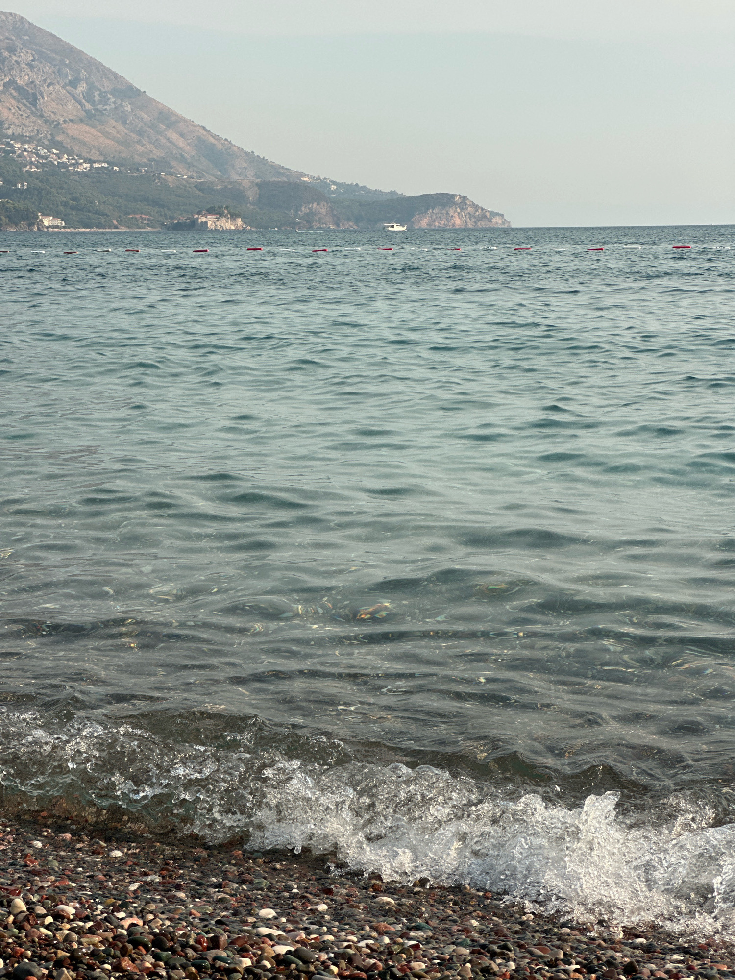 The image depicts a coastal scene with a pebble beach in the foreground and the sea extending into the background. A gentle wave is breaking on the shore, creating white foam and splashes. The water is clear enough to see the submerged pebbles. In the middle ground, a line of buoys floats on the water. Further in the distance, a mountainous coastline is visible, with some buildings and greenery clinging to the slopes. A small white boat is also present on the water, near the coastline. The sky above is a pale, hazy blue, suggesting an overcast or lightly foggy day. The location is identified as Boreti, Montenegro.