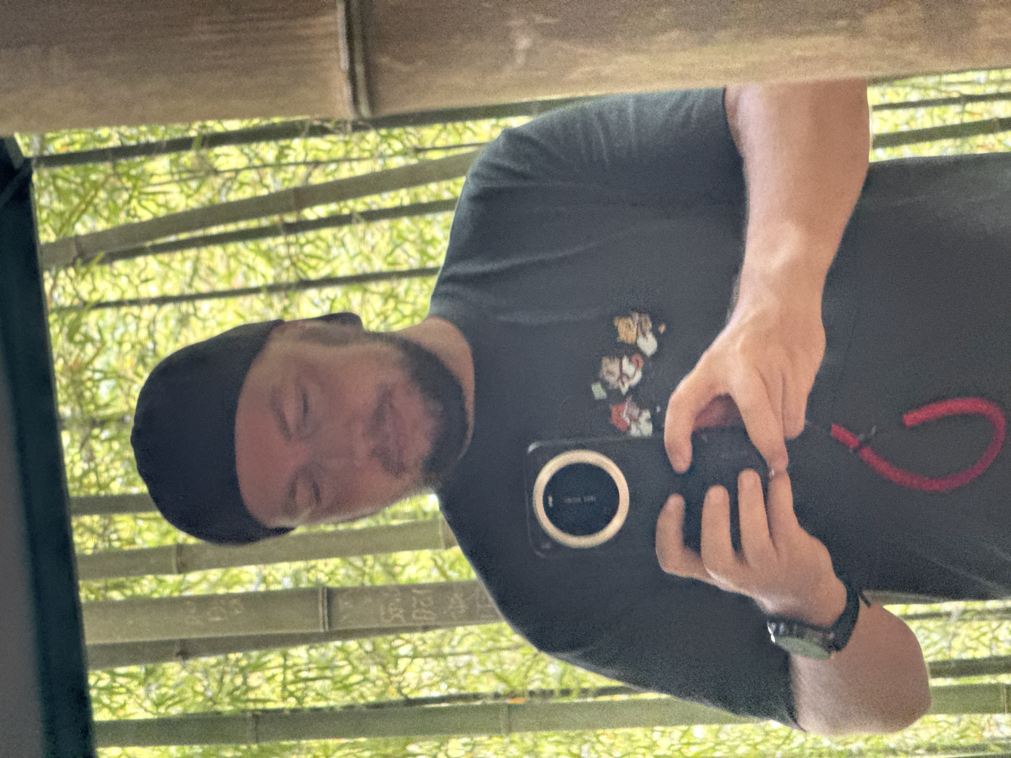 A man wearing a black t-shirt and a black cap is taking a selfie with his smartphone in front of a bamboo fence. The environment suggests Púng Luông, Vietnam. The phone has a red lanyard attached. The man is wearing a watch on his left wrist. The background is filled with lush green foliage. The image seems to be taken from a low angle looking up, possibly reflecting off a surface.