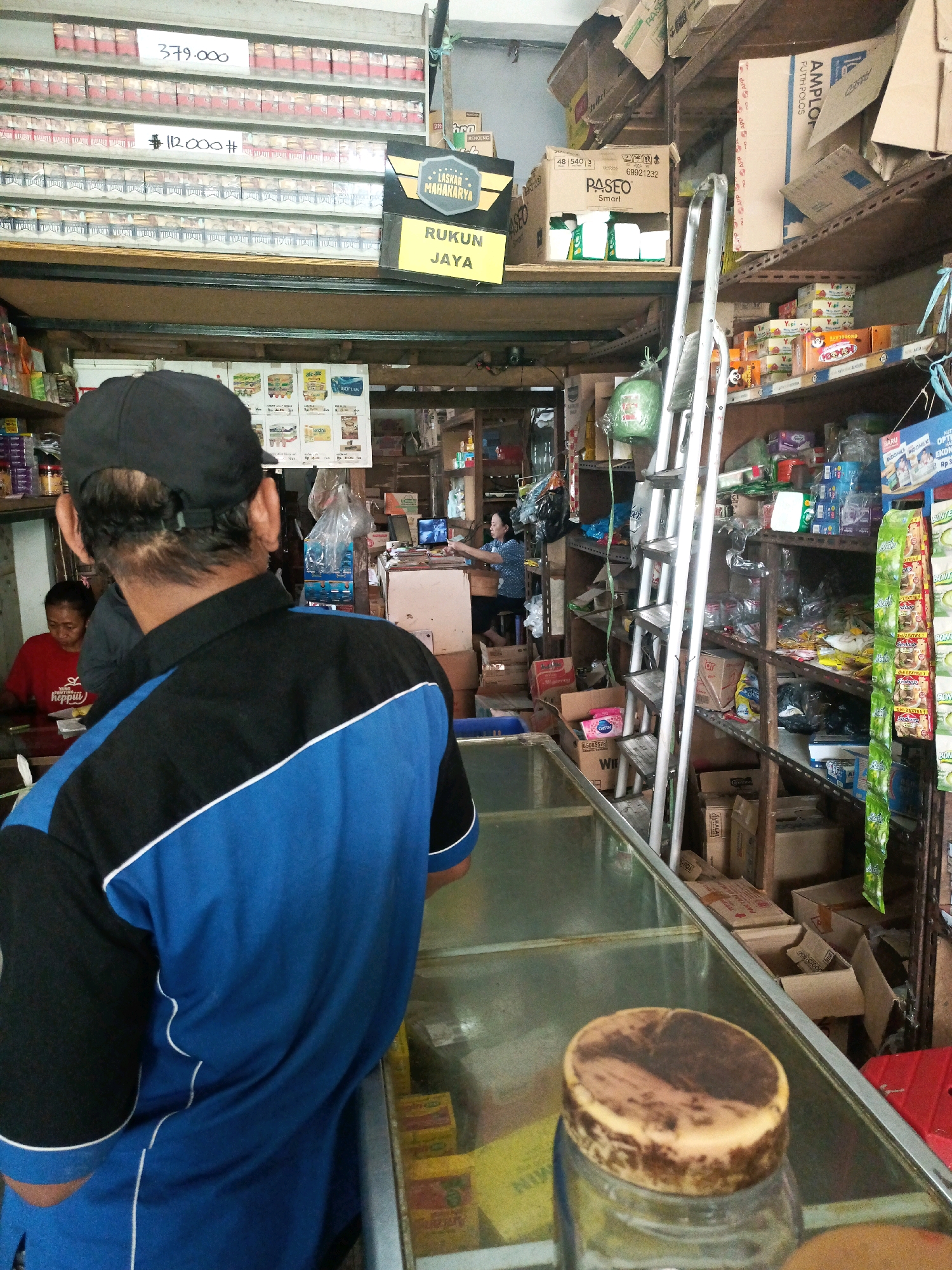 The image depicts the interior of a small convenience store, likely in Malang, Indonesia, given the presence of Indonesian text on signage and products. The store appears to be well-stocked, with shelves filled with various goods, including cigarettes, packaged food items, and household products.
In the foreground, a person wearing a blue and black polo shirt with white trim is seen from behind, facing away from the camera. Their back is to the viewer as they stand behind a glass display counter. Their head is covered by a black baseball cap.
Behind the counter, to the left, another person in a red t-shirt is visible, facing the camera. They appear to be a store employee or customer. Further back, a woman is seated at a desk with a laptop, possibly managing the store's operations.
The store's shelves are packed with merchandise. Cigarette packs are prominently displayed on upper shelves, some with price tags indicating "379.000" and "112.000". Other shelves contain a variety of packaged goods, including what appear to be food items like instant noodles and snacks, as well as cleaning supplies and toiletries. Some shelves are also stocked with boxes and plastic-wrapped items.
A tall, silver ladder leans against a shelf unit, suggesting that higher items are accessible. Several cardboard boxes are stacked on the floor and on shelves. A green plastic bag containing an unknown item is hanging from a shelf.
The overall impression is of a busy, albeit somewhat cluttered, local shop. The lighting suggests it's daytime, with overhead fluorescent lights providing illumination. The environment is functional, with a focus on product display and storage.
