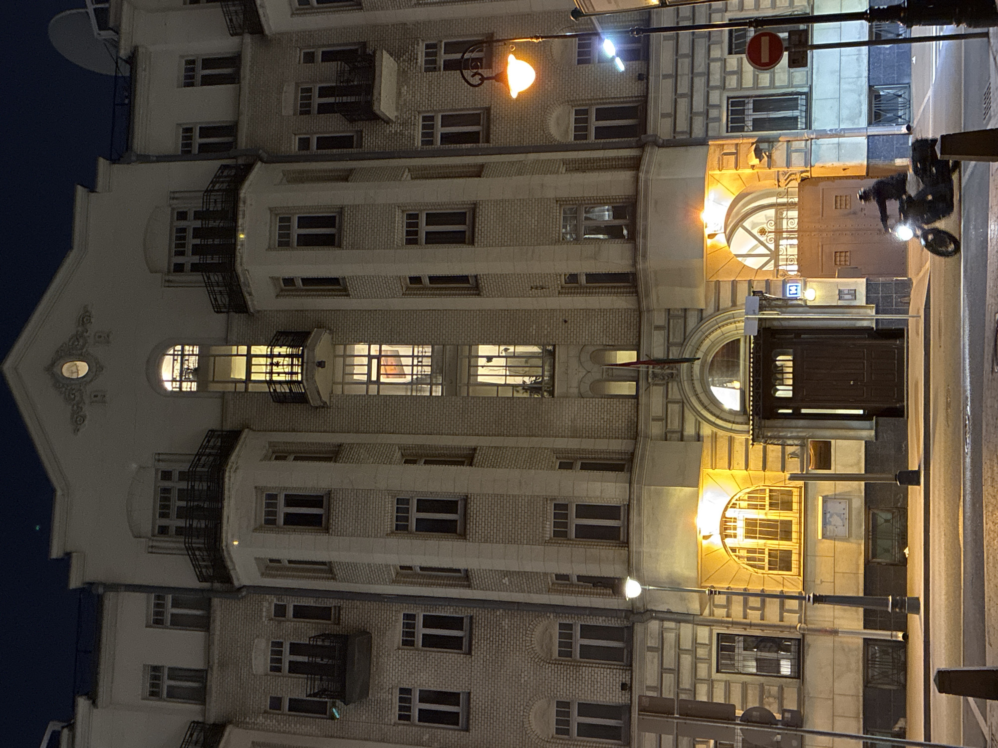 The image shows a nighttime urban scene in Moscow, Russia. The primary subject is the facade of a multi-story brick building. Architectural details include arched windows, balconies, and decorative molding. Streetlights illuminate the building and the street. A person is riding a scooter on the street. Street signs are visible. The overall lighting is dim.
