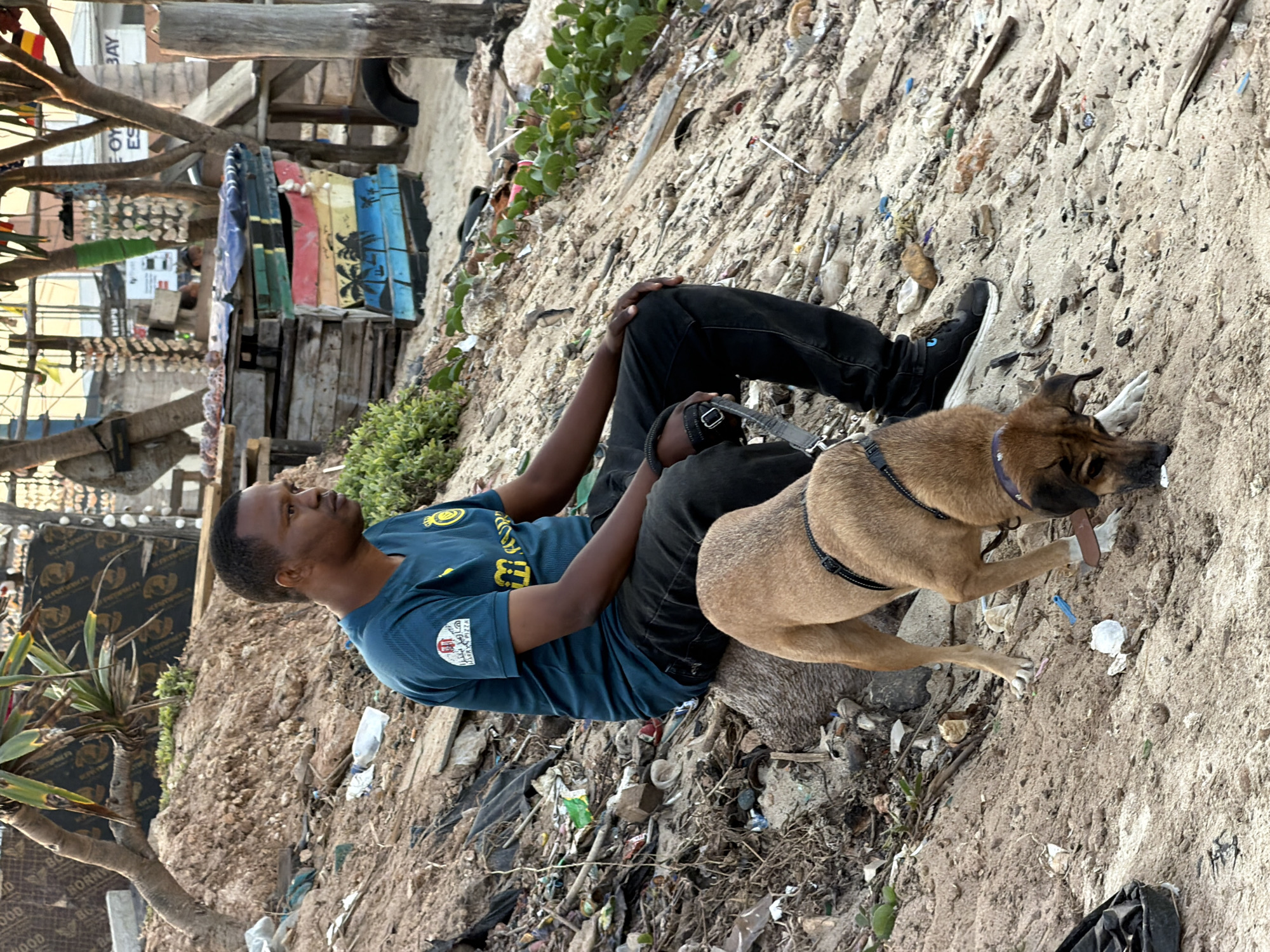 A man sits on a sandy embankment with a medium-sized brown dog. The man is wearing a dark teal t-shirt with yellow lettering and a logo, and black jeans. The dog is wearing a black harness. The embankment is covered in debris, including plastic bottles, bags, and wood fragments. In the background, there is a weathered wooden structure painted with colorful horizontal stripes and a scene of palm trees. Behind that, a sign reads "E-OY ES BAY". The scene appears to be a beach area, likely along the coast of Dar es Salaam, Tanzania, characterized by its sandy terrain and coastal vegetation. The overall impression is one of a relaxed, perhaps informal, setting with signs of human activity and waste.